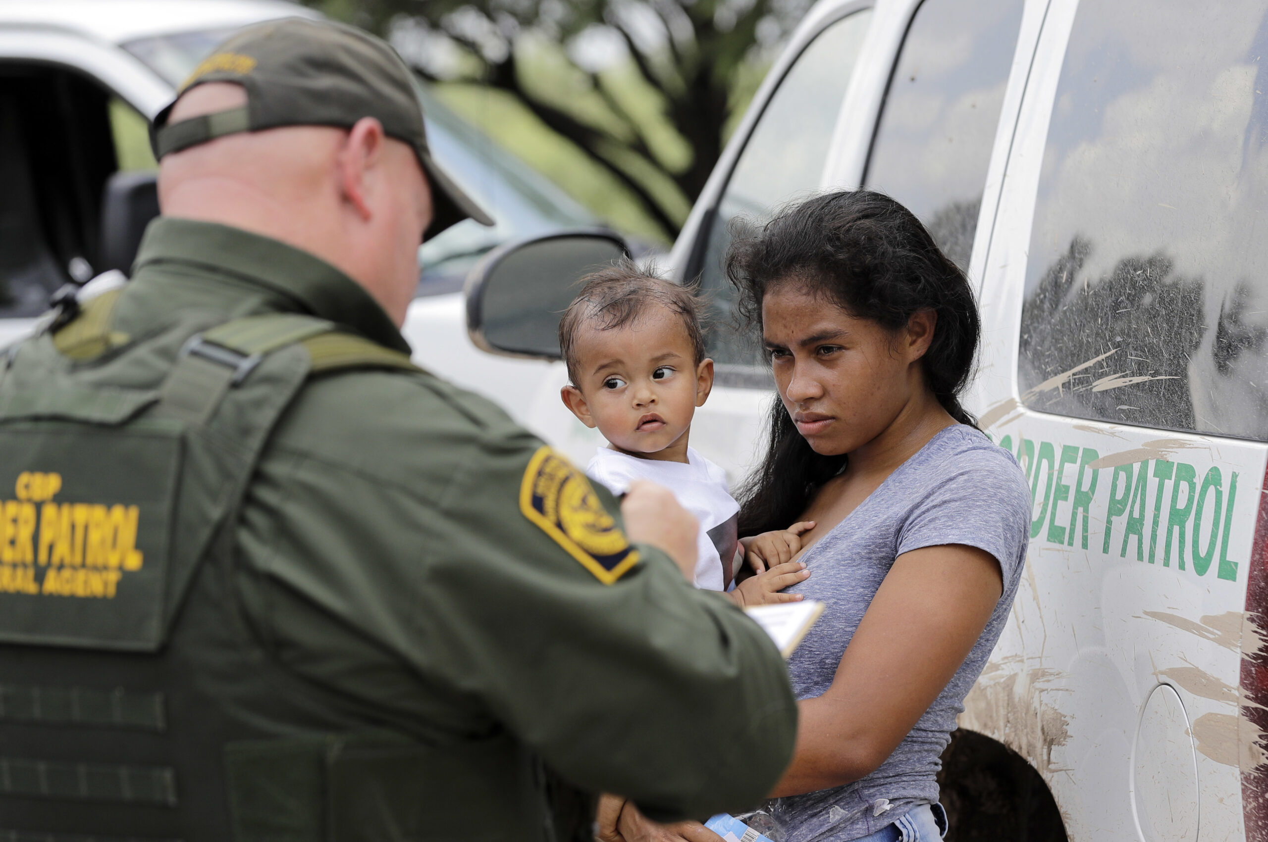 A border patrol officer in a green uniform interviews a Latina migrant holding a baby, while both are standing in front of a U.S. Customs and Border Patrol van.