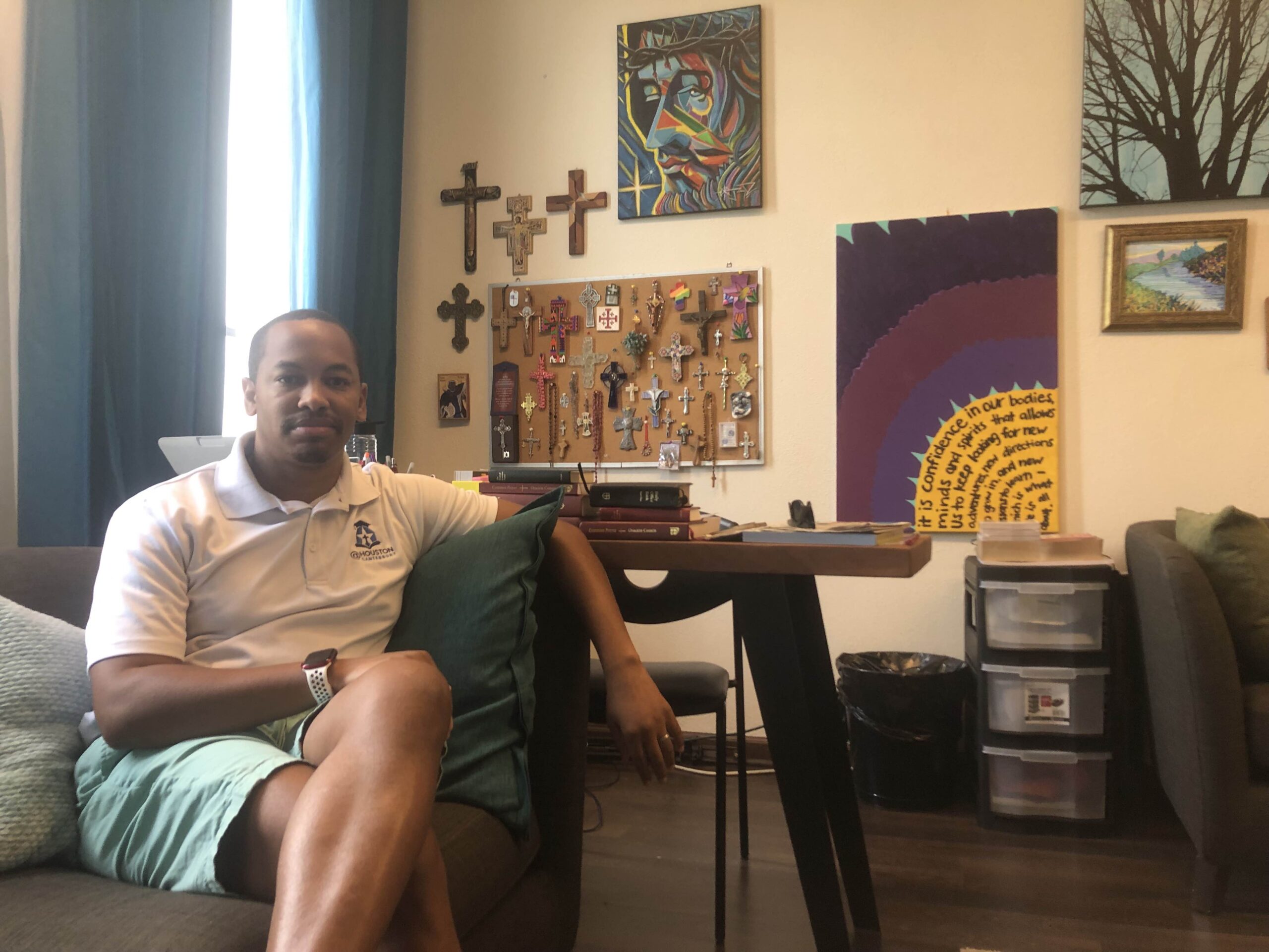 Charles Graves, a Black man in a polo shirt and shorts, sits in a comfortable looking office, with artwork and a collection of crosses in the background.