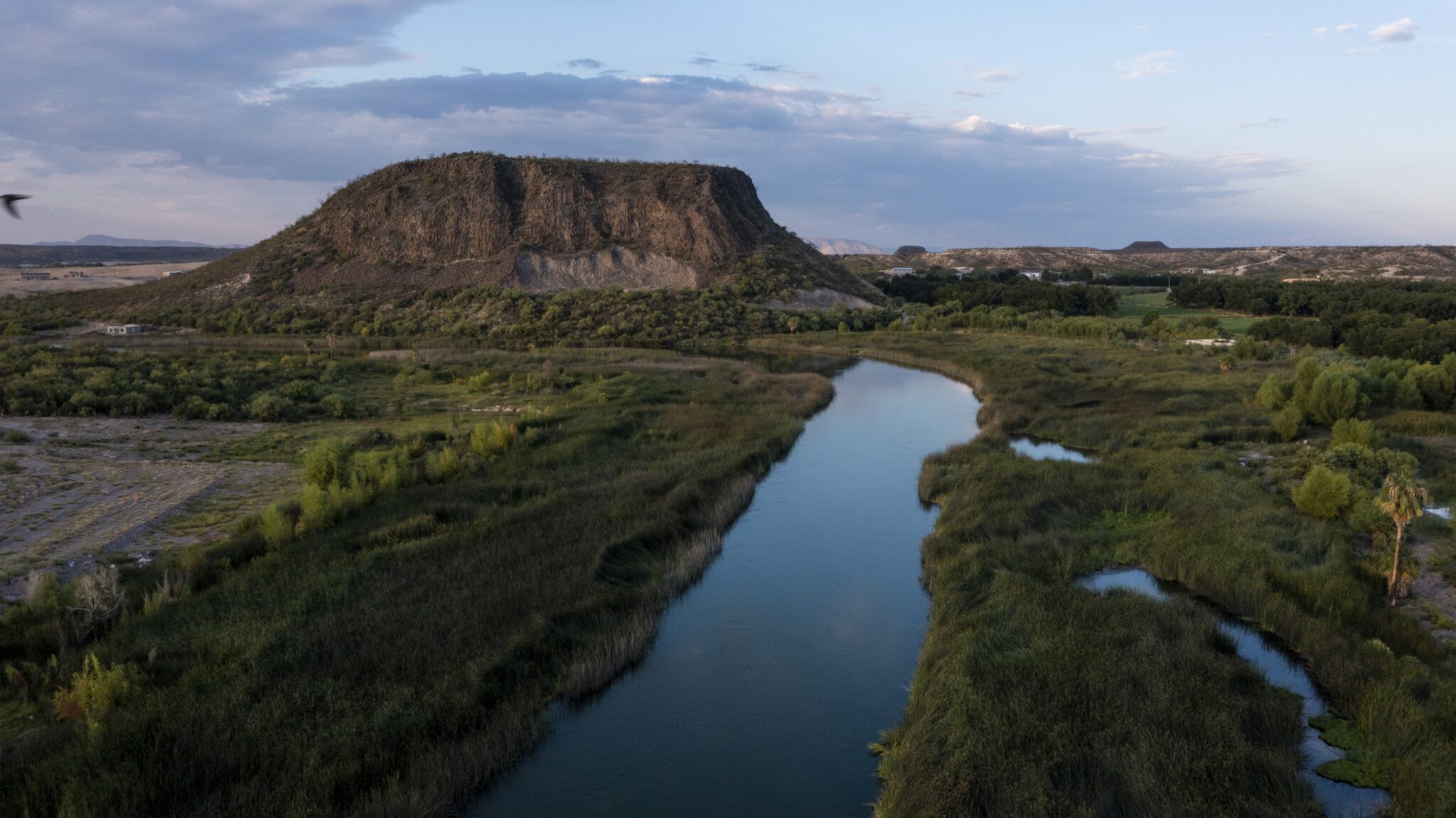 A river winds through a lush looking desert landscape, past a lone mountain.