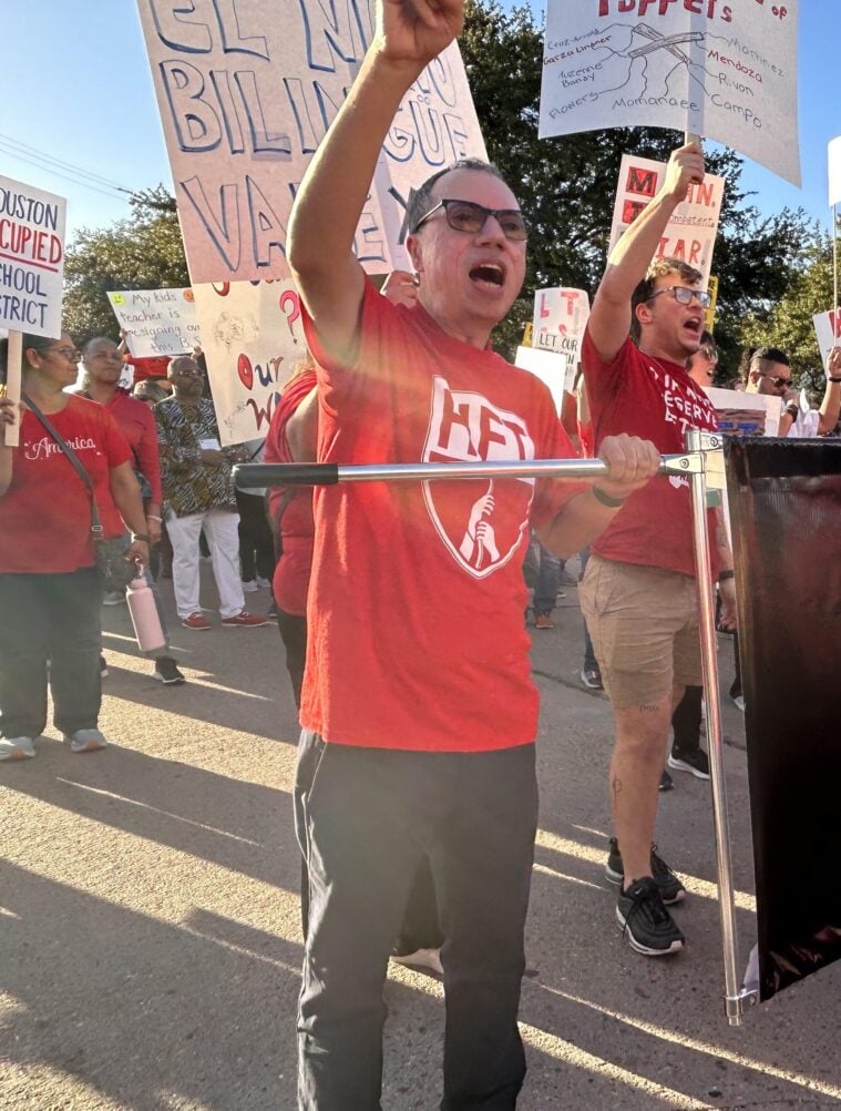 A man in a red Texas Houston teachers' union t-shirt marches with hundreds of others in the streets, one fist upraised.