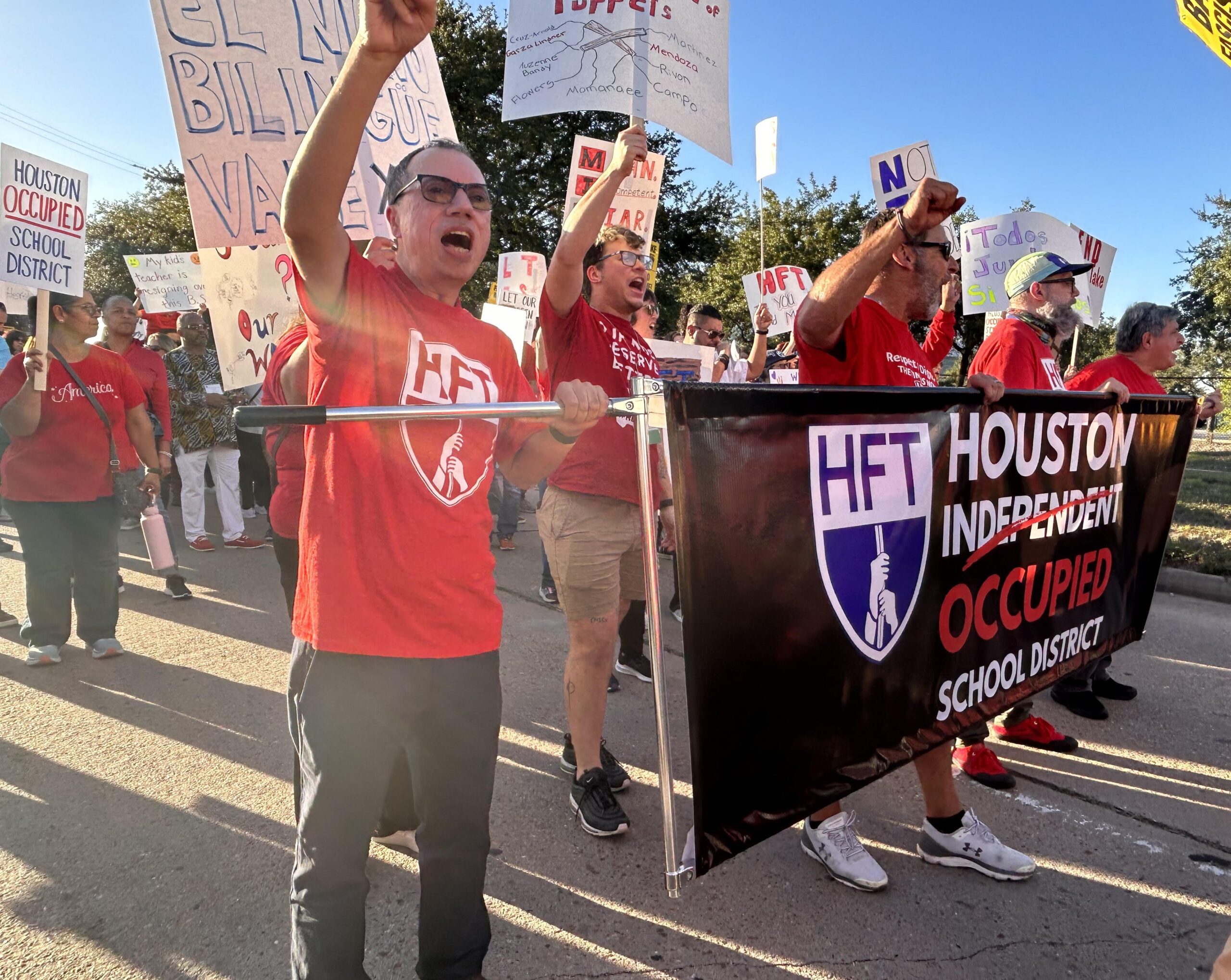 Men in red t-shirts march at the head of a crowd with their fists upraised. They are holding a Houston teachers union banner.