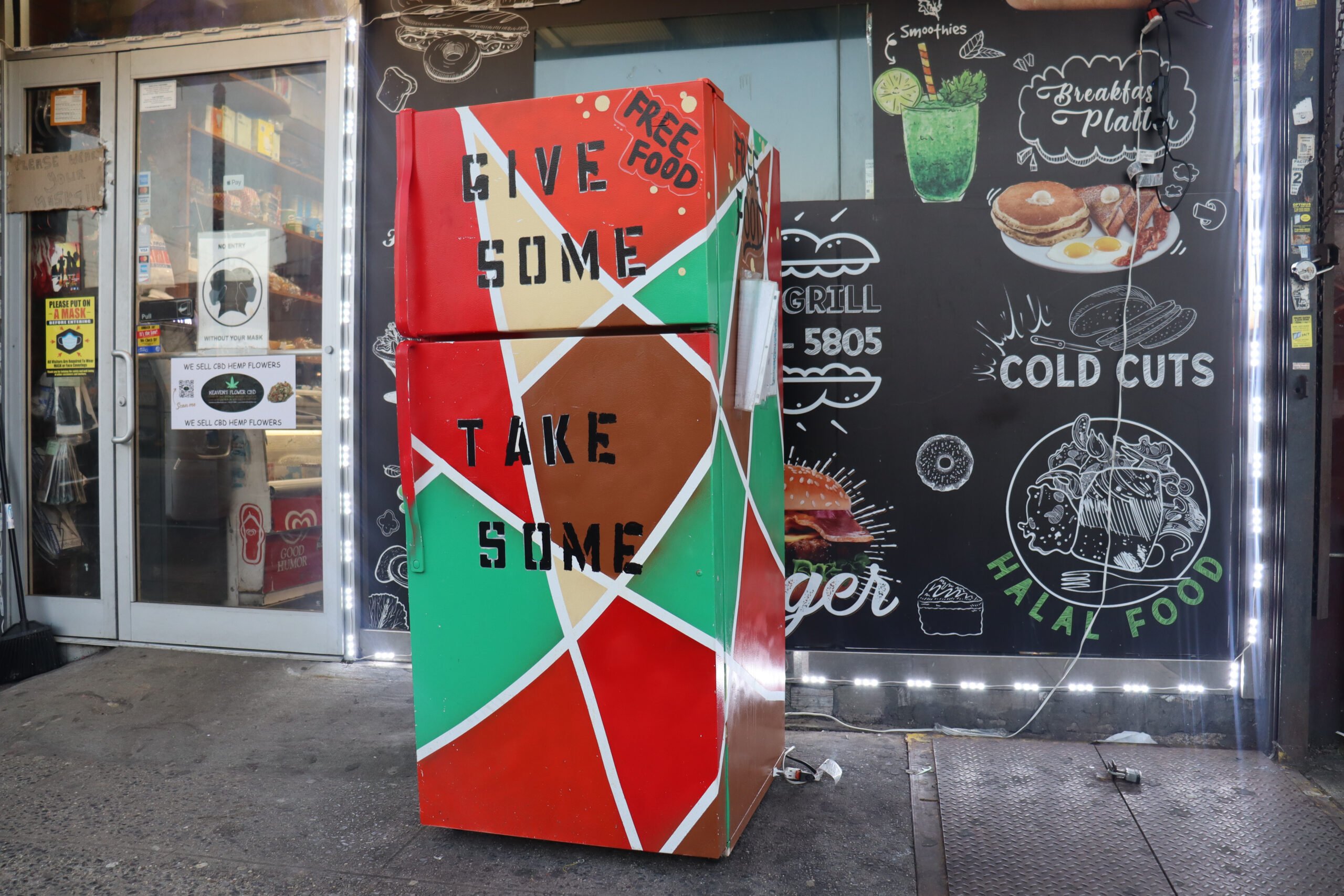 A community fridge painted red, green, brown and yellow in a mosaic-like pattern reads "Free food. Give some, take some." It appears to be outside a deli or restaurant.