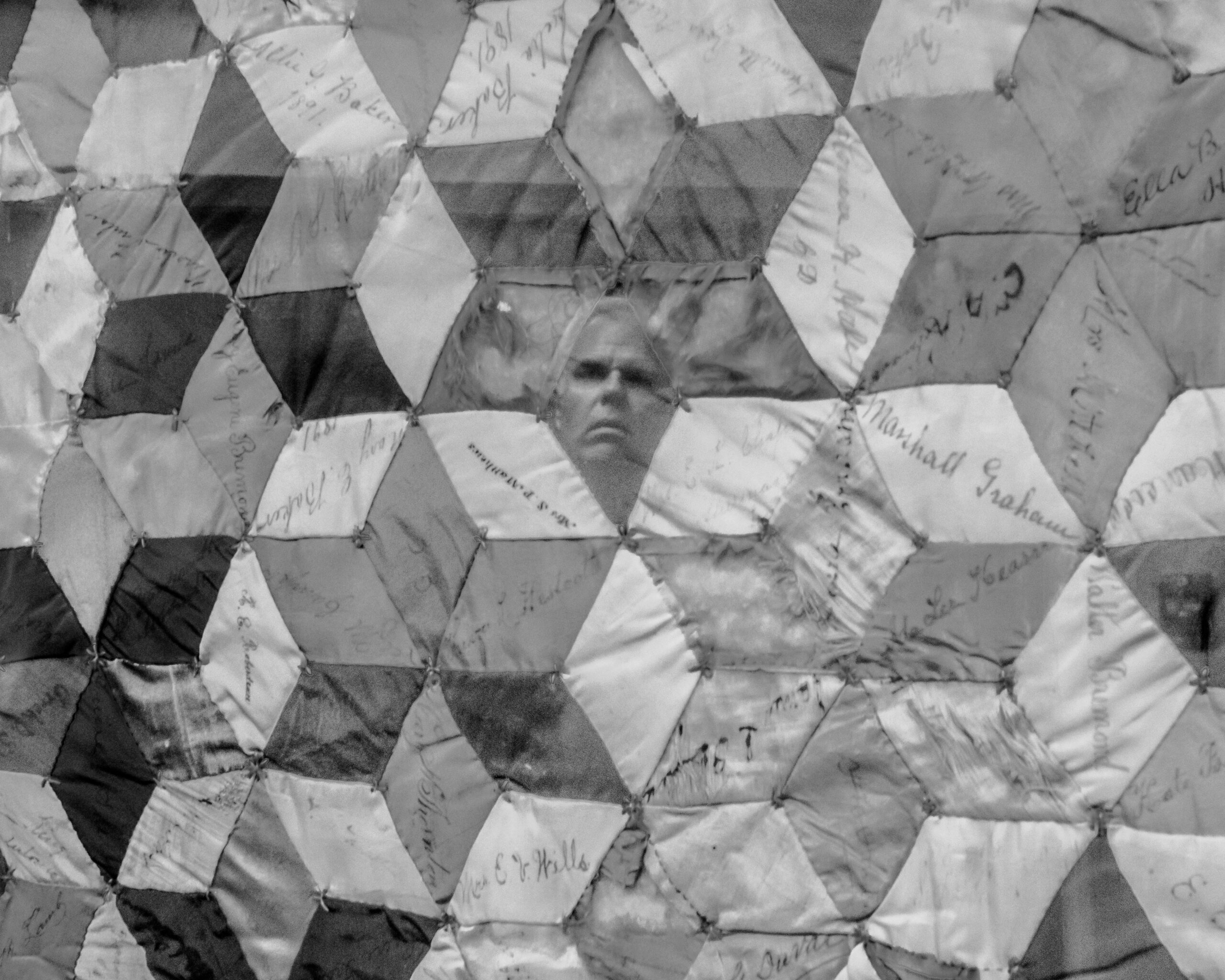 The face of Rob Faubion, an older white man, can be seen in the reflection over the glass frame of an antique quilt, which is made from interlocking diamond shapes, many of them signed by donors to the Confederate Women's Home.