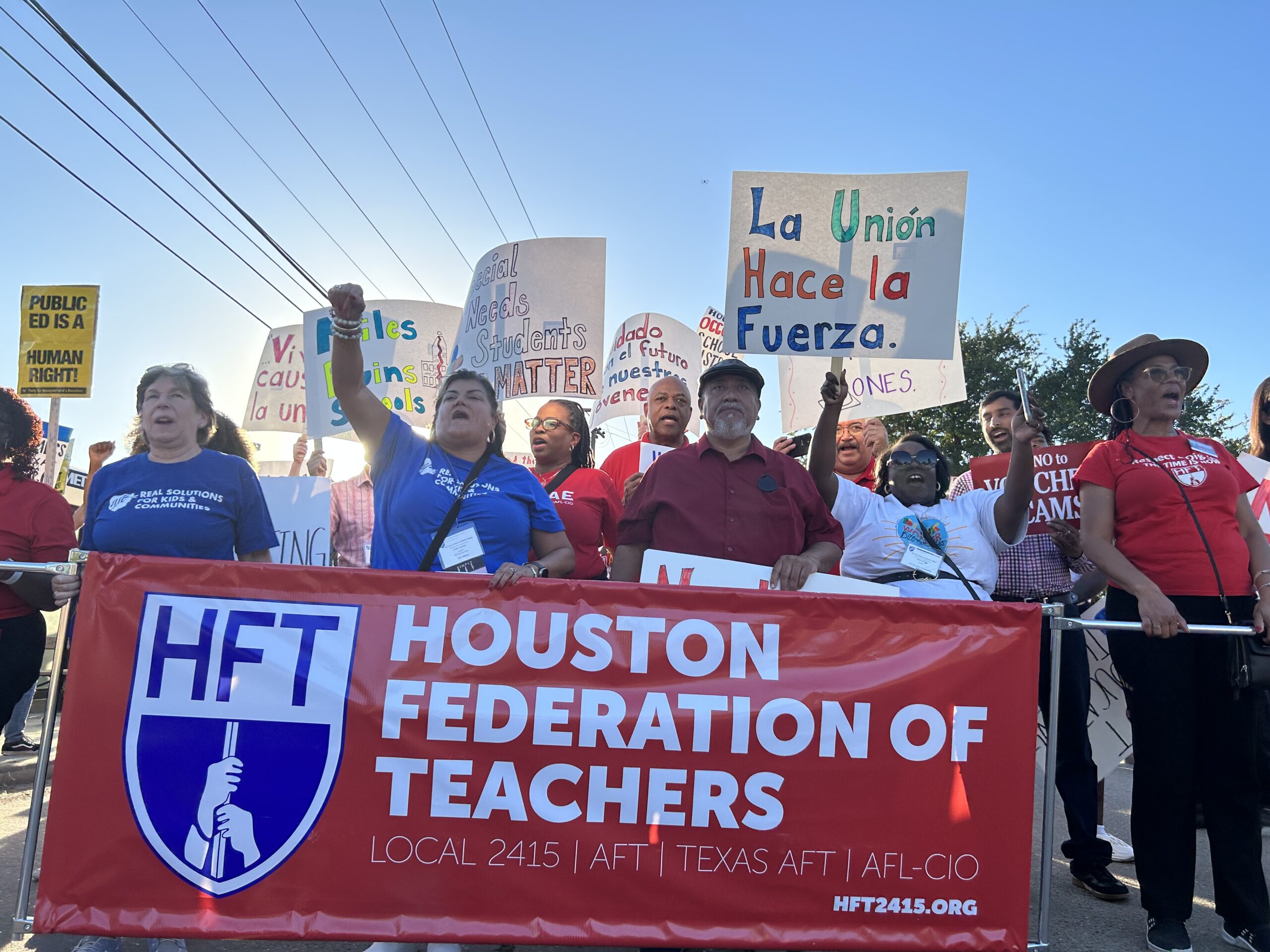 A diverse group of men and women, many in red union t-shirts, with several in front holding a large Houston Federation of Teachers banner, march in the street.