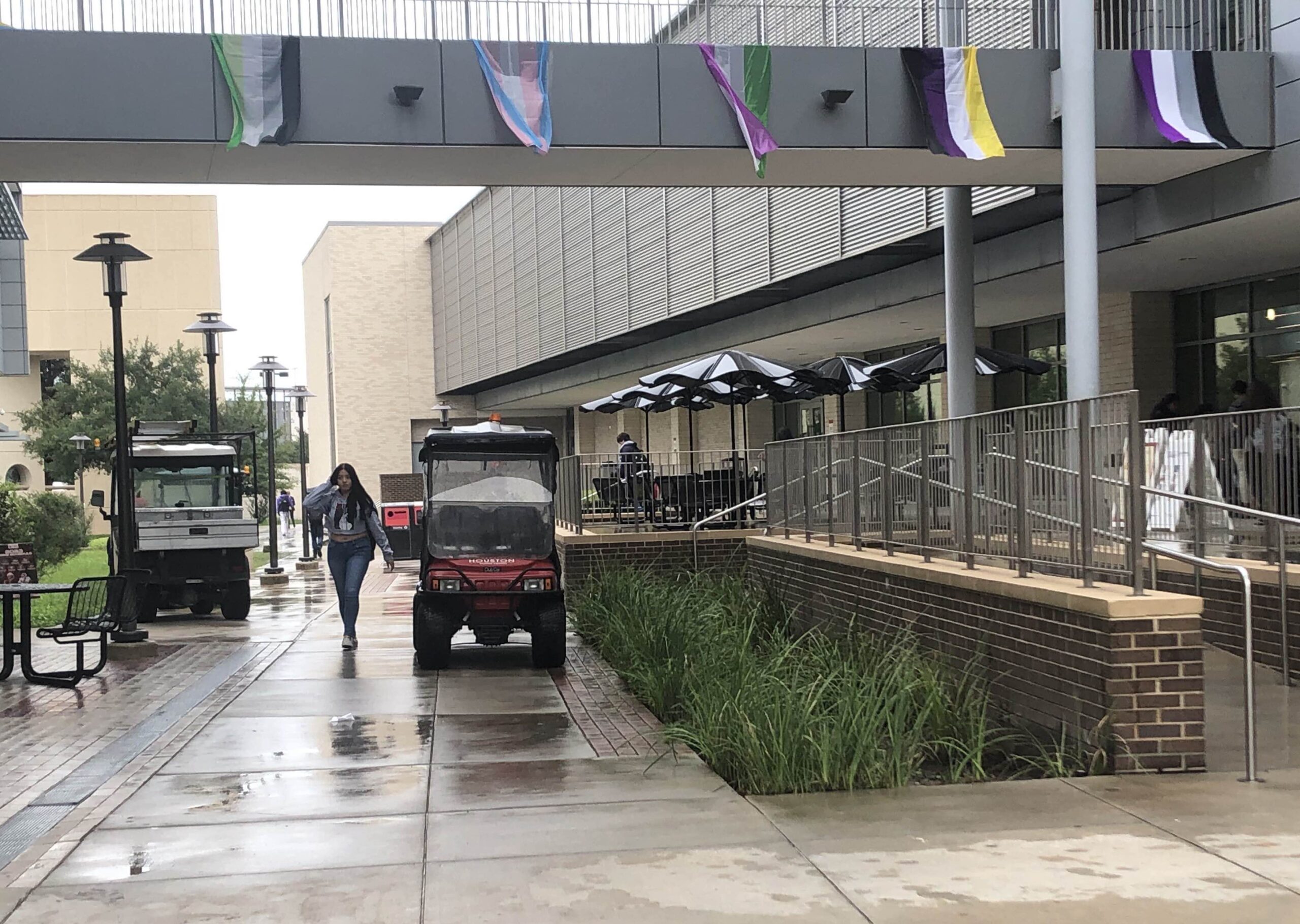 Various LGBTQ+ Pride flags hang from an elevated walkway above the rain-soaked campus of the University of Houston. A few students walk outside while faculty or staff drives past in covered golf carts.