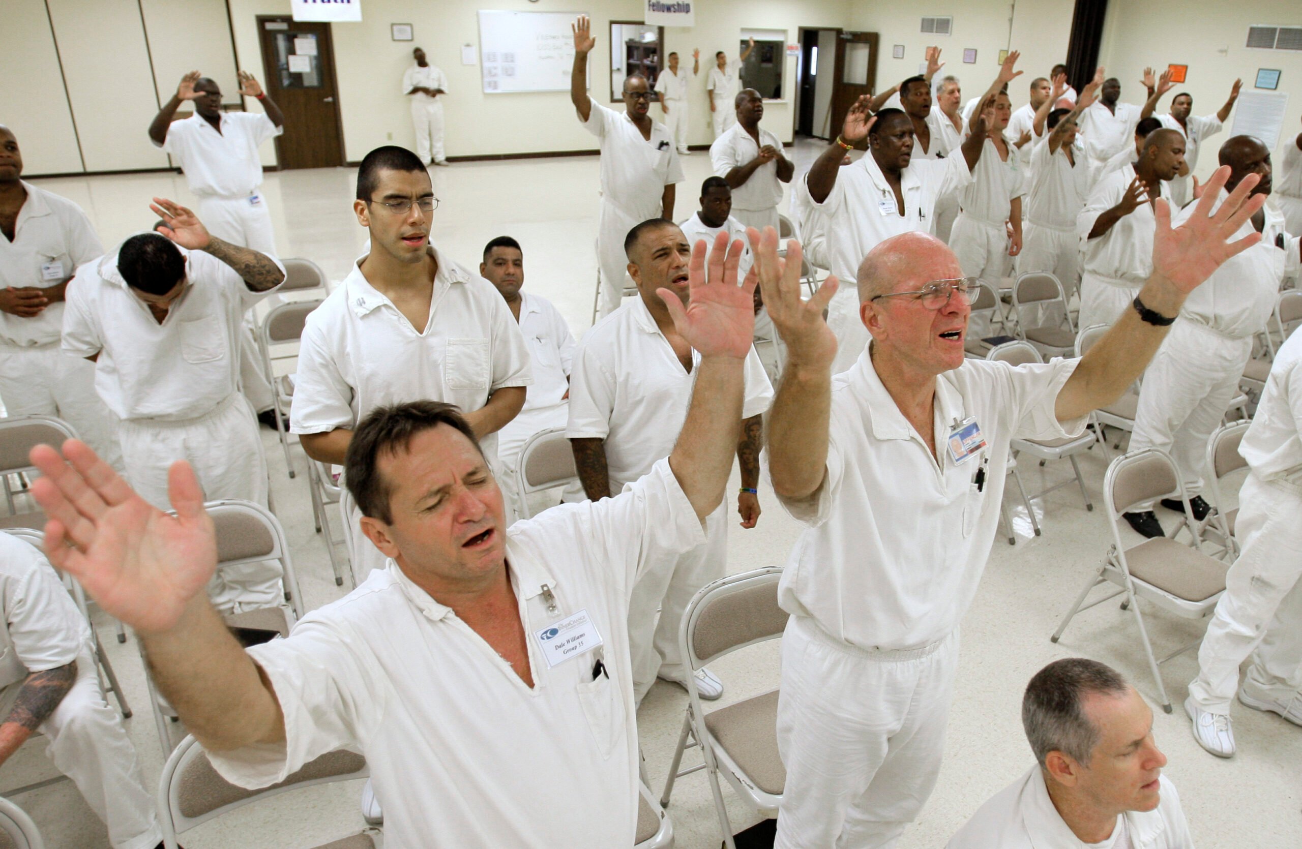 Male inmates in white prison uniforms stand near rows of folding chairs. Their arms are raised with expressions of praise, worship and focus on their faces. The crowd looks diverse with both Black, white and Latino worshipers.