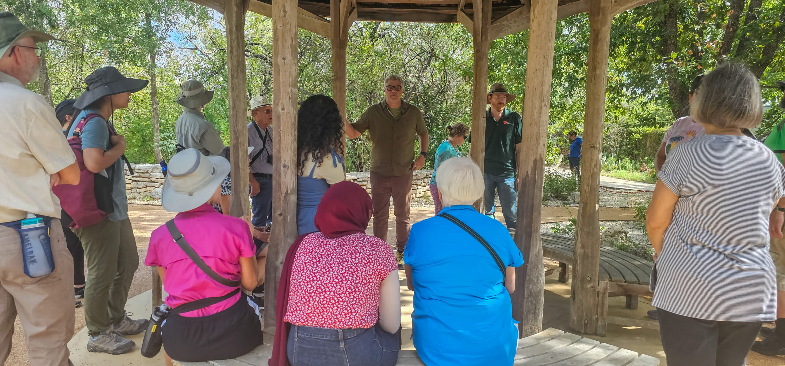 A group of people are gathered under an outdoor gazebo, listening intently to a discussion about Texas native plants led by a man in a green shirt.