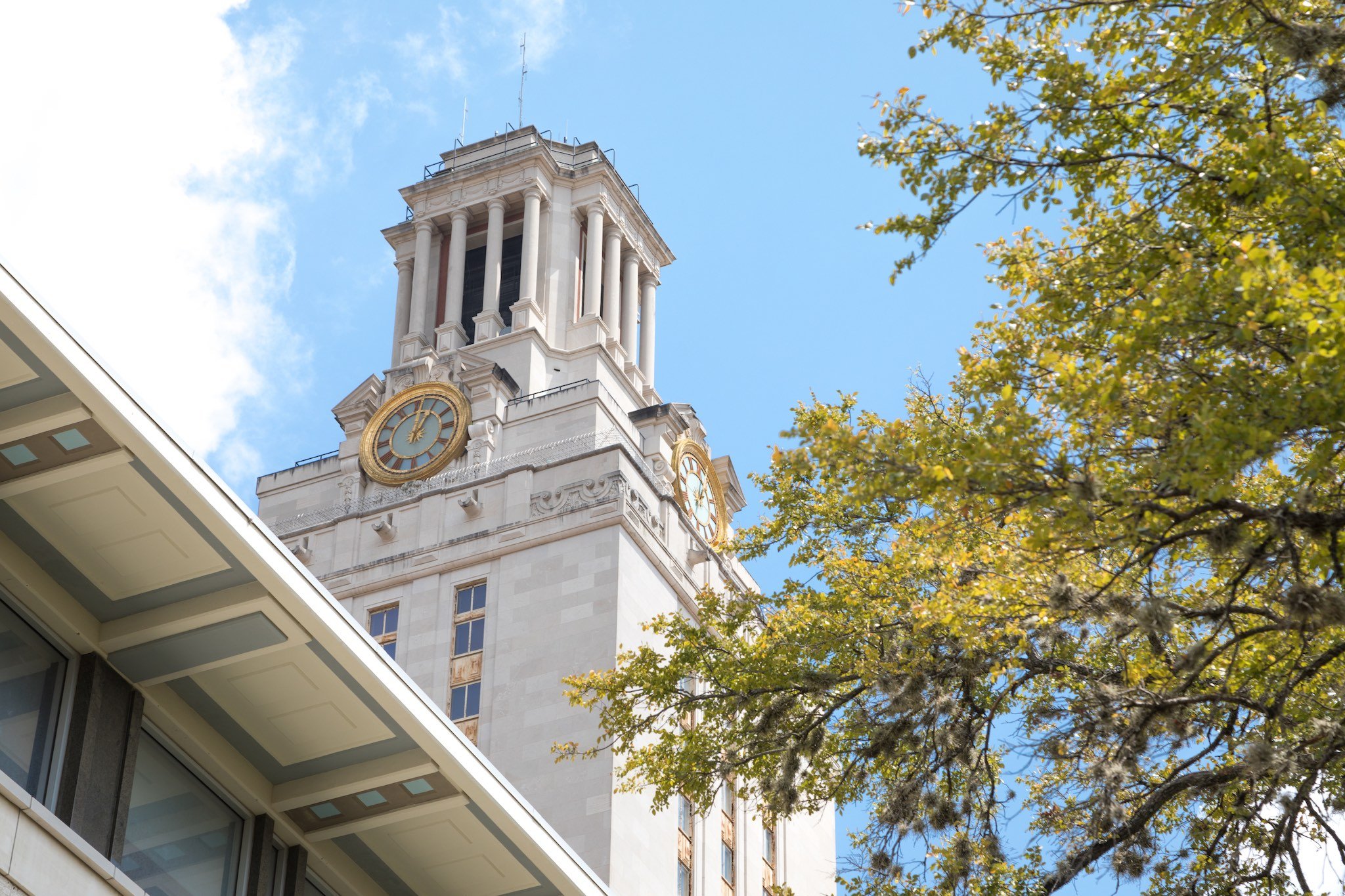 A low angle view of the UT Clock Tower with trees in the foreground at the University of Texas at Austin, under a partly cloudy blue sky.