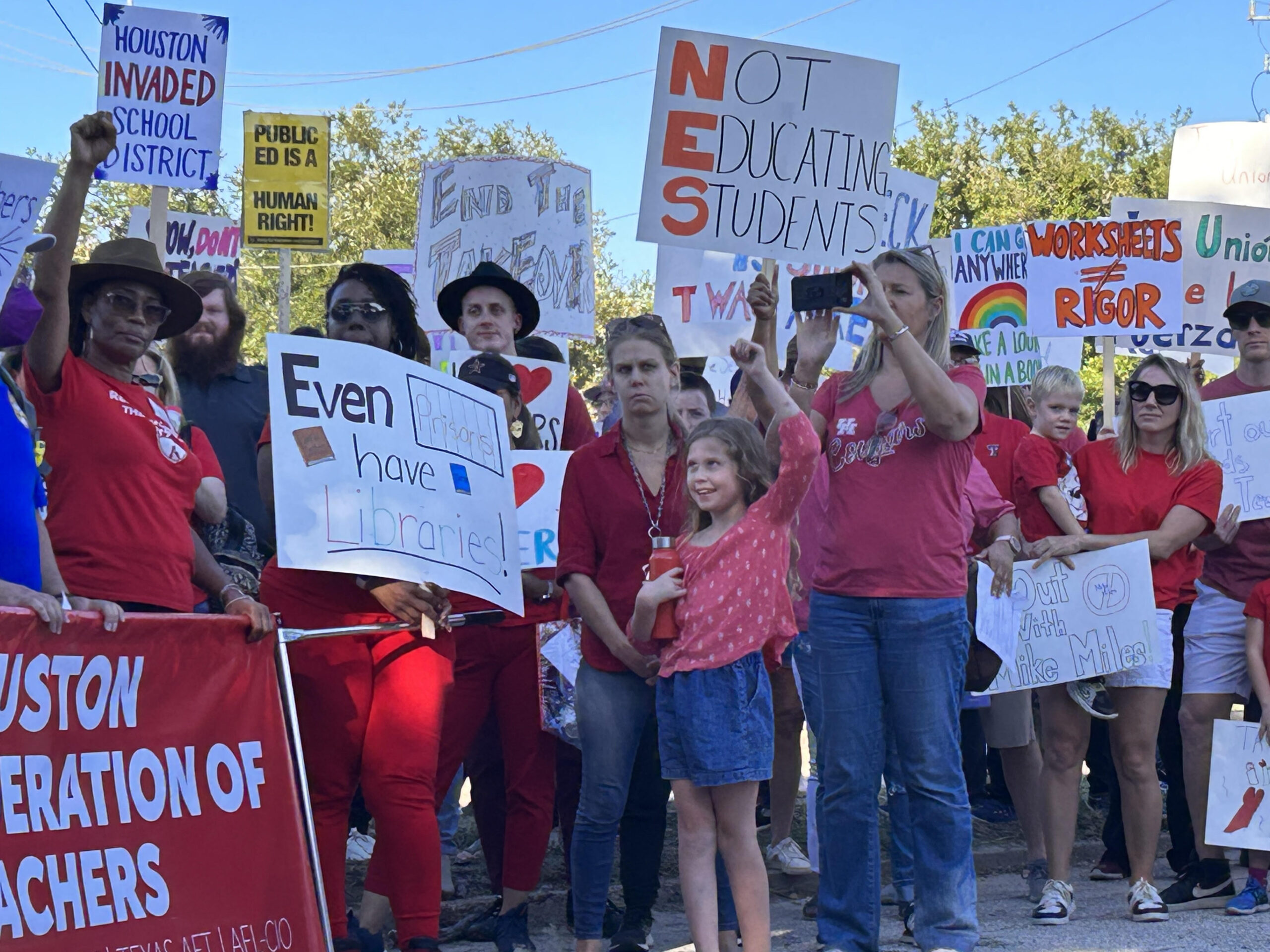 A diverse crowd poses during a protest press conference, many in red t-shirts or holding signs opposing the state takeover of Houston ISD by Mike Miles.