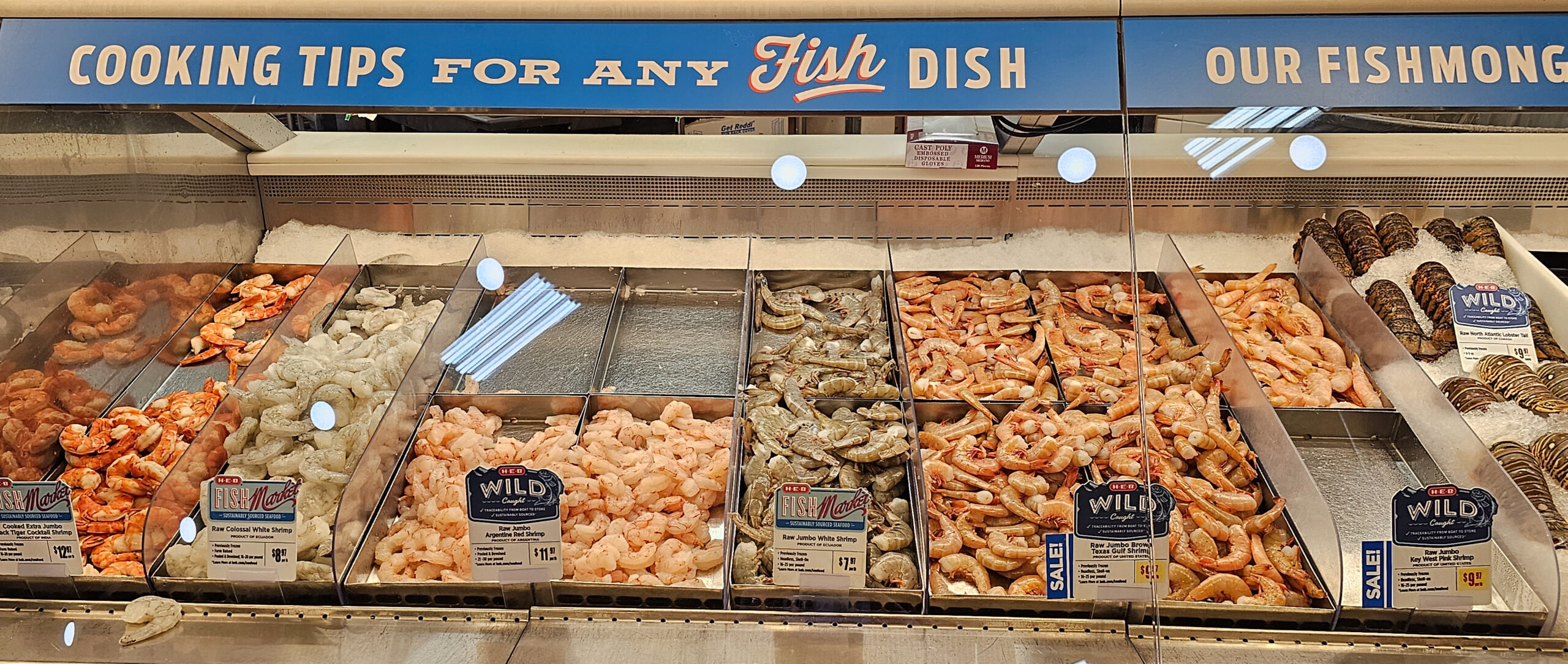 A refrigerated grocery store display of shrimp and seafood, with a sign reading Our Fishmongers have Cooking Tips for Any Fish Dish.