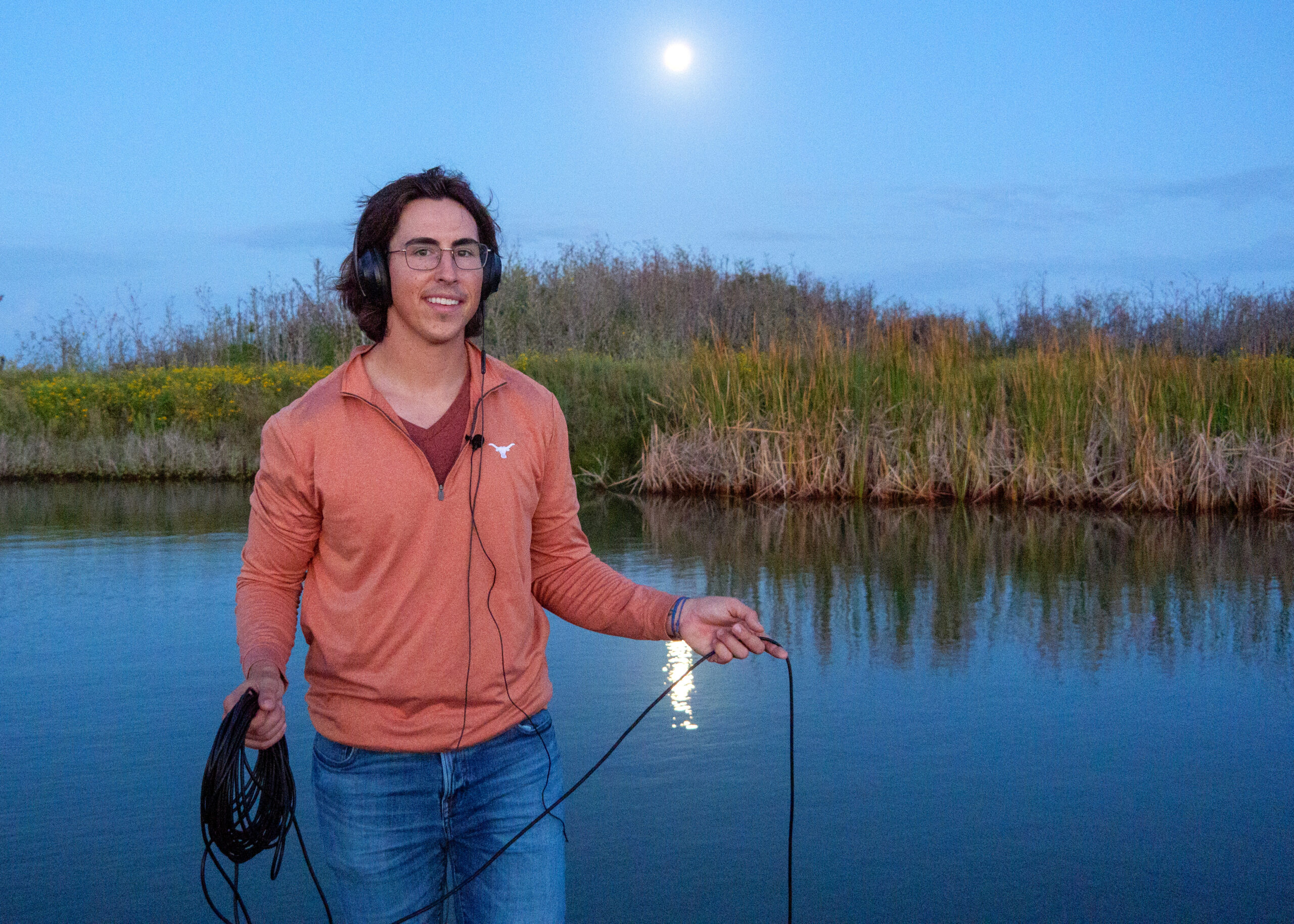 Philip Souza poses by the bay of Port Aransas with his fish sound recording gear. He's smiling, dressed in blue jeans and a burnt orange UT hoodie.