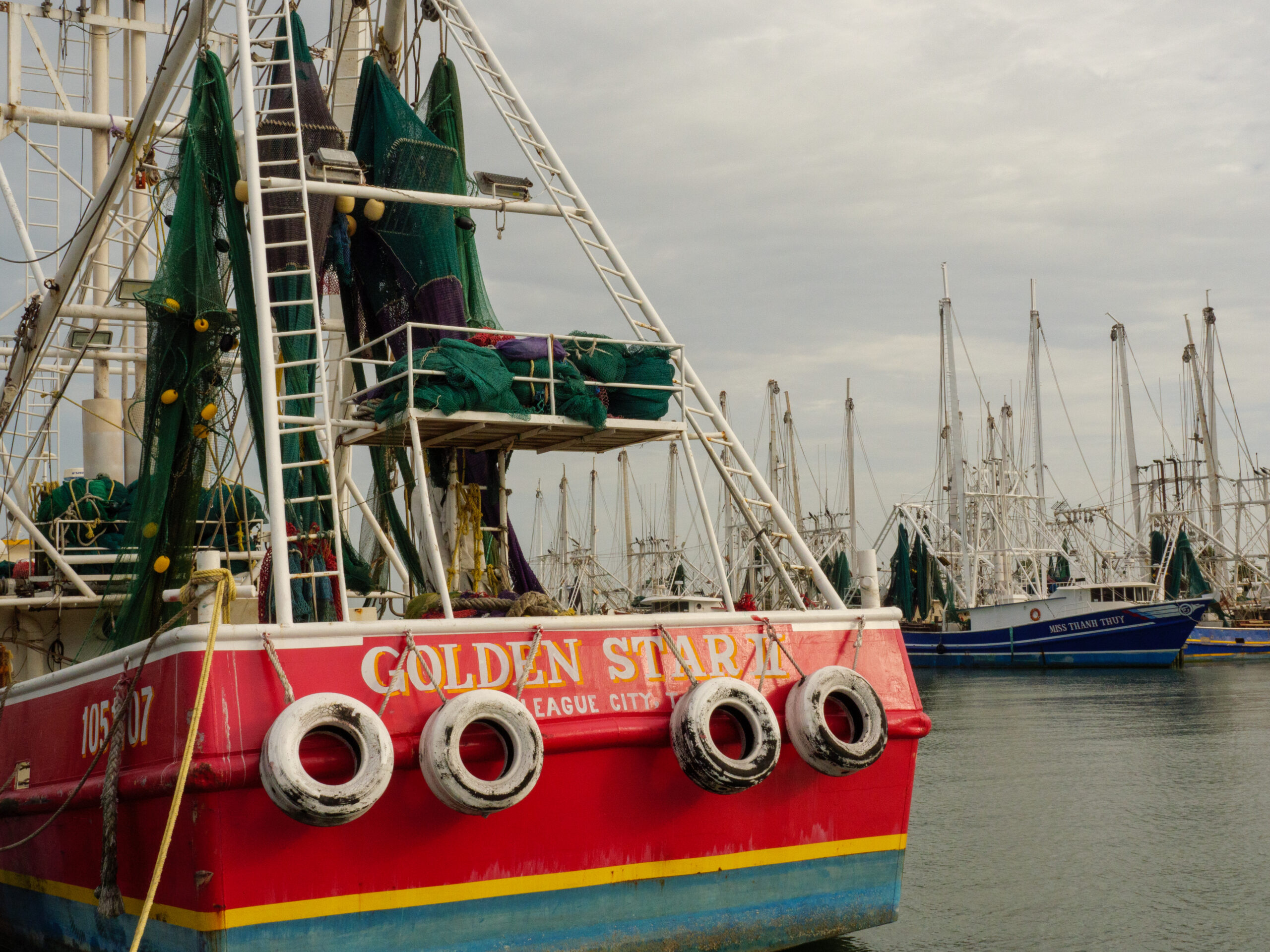 A red ship with its name in white, also declaring the boat is from League City. Green shrimp nets hang from the masts. Many other shrimp boats are visible in the densely packed marina.