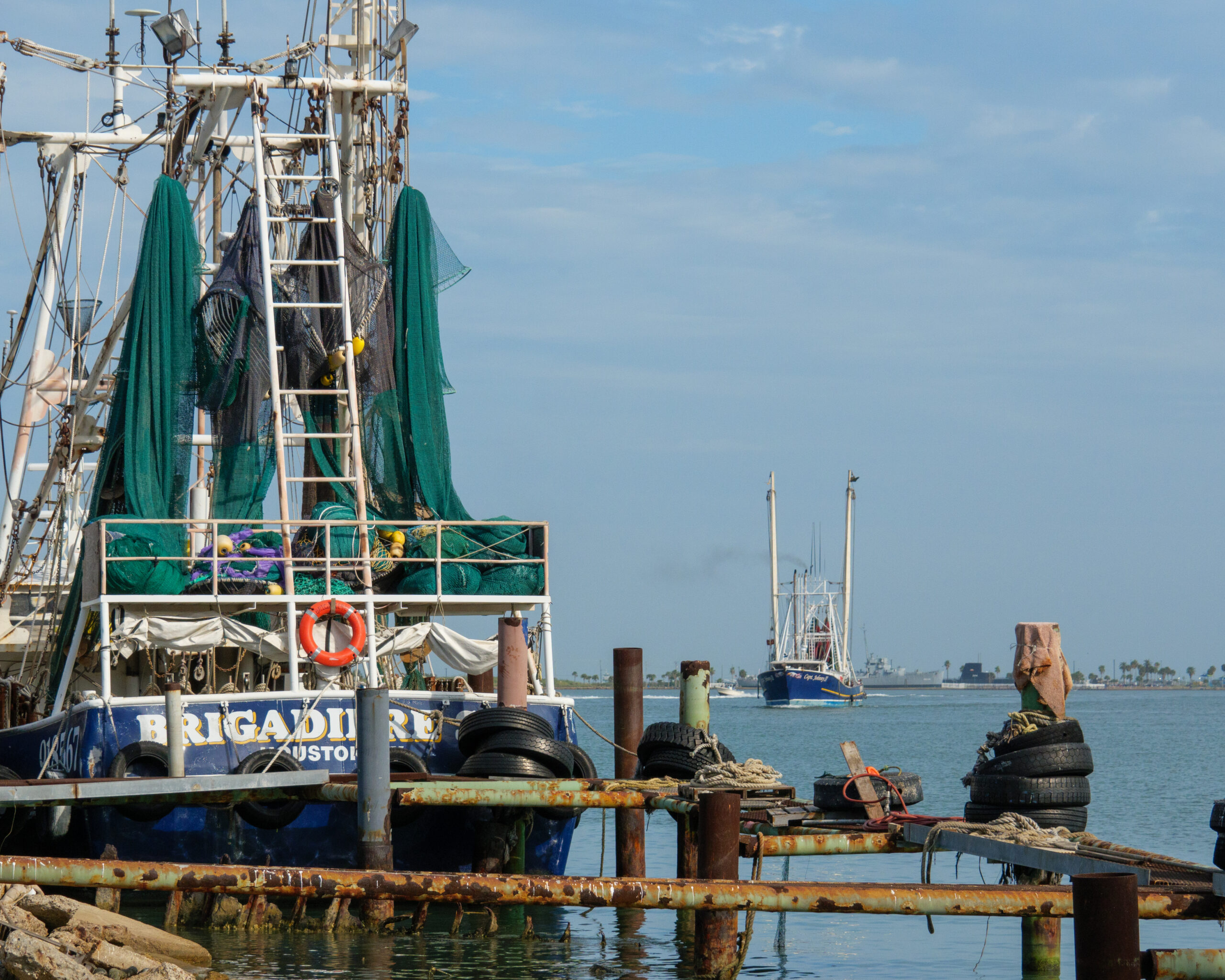 The stern of the fishing boat in the foreground declares it to be from Houston. Green nets hang from the rigging. In the background, another ship returns from sea.