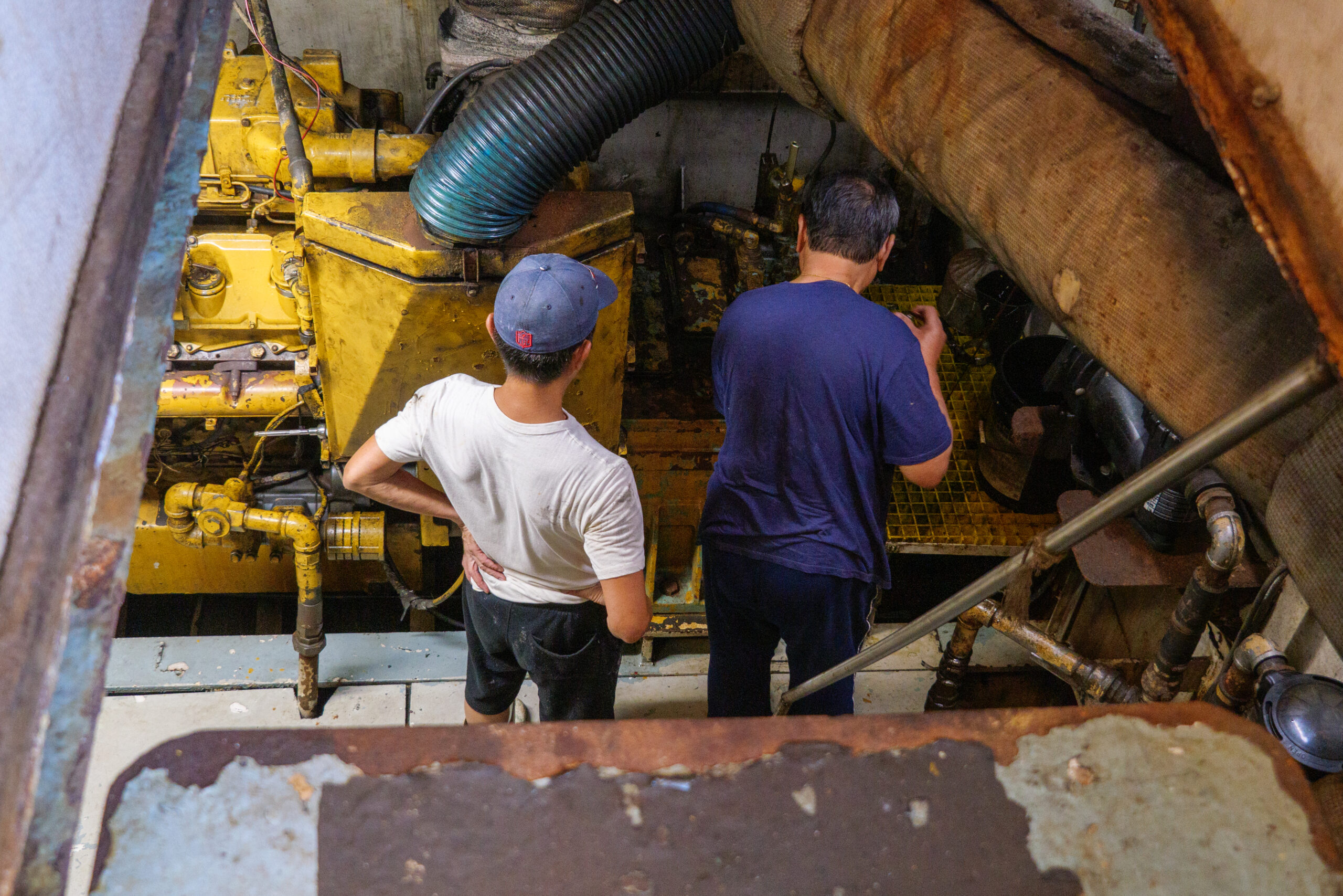 A view of the engine hold from the rusted, weathered metal of the deck. Seen from behind, the two Vietnamese men tend to the engine. The man to the left is wearing a blue ballcap, white tee and black shorts, while the other is in a blue tee, and striped track pants.