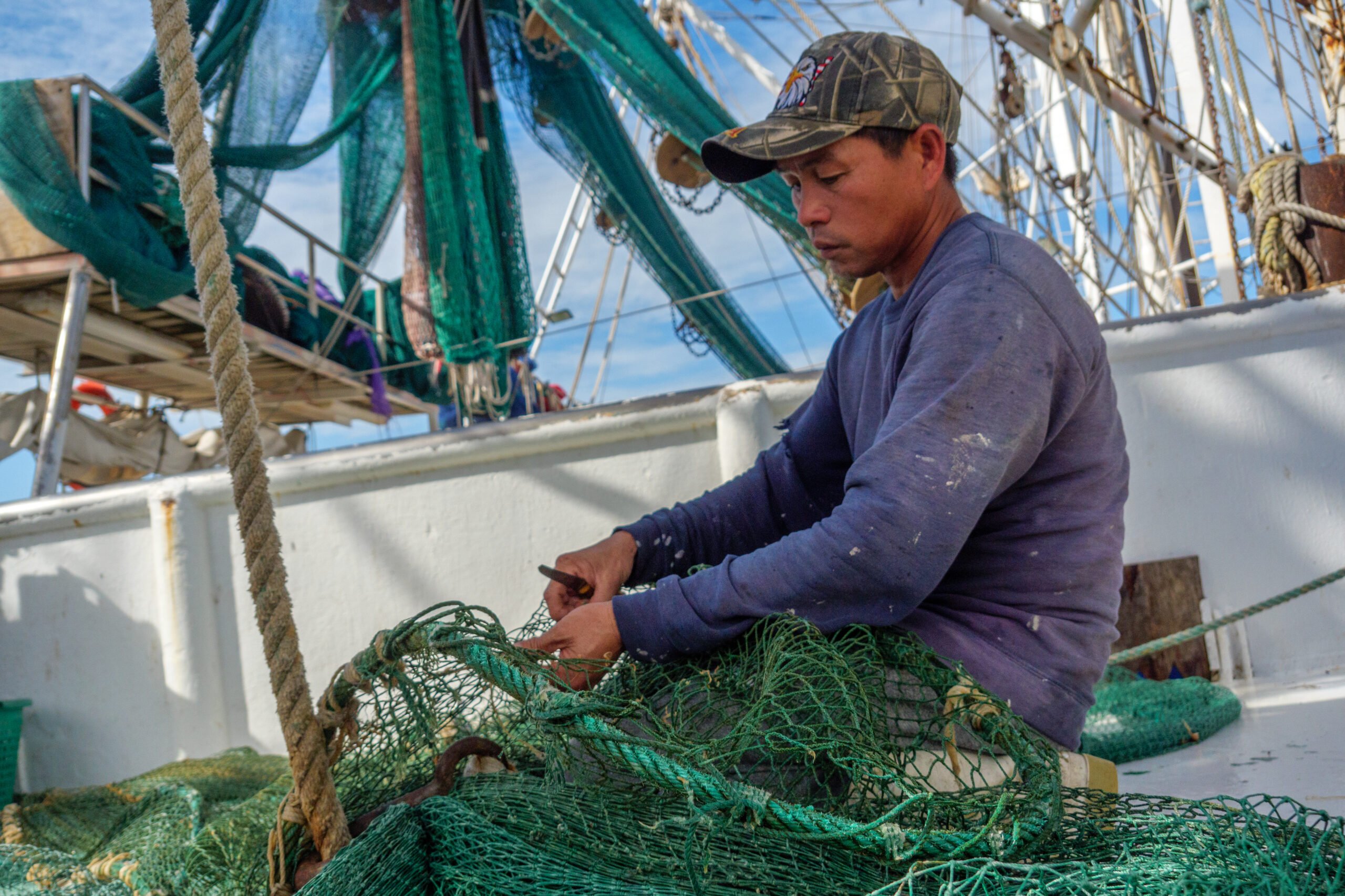 A Vietnamese man works on bright green shrimp nets aboard the slanted deck of a shrimp boat at sea.