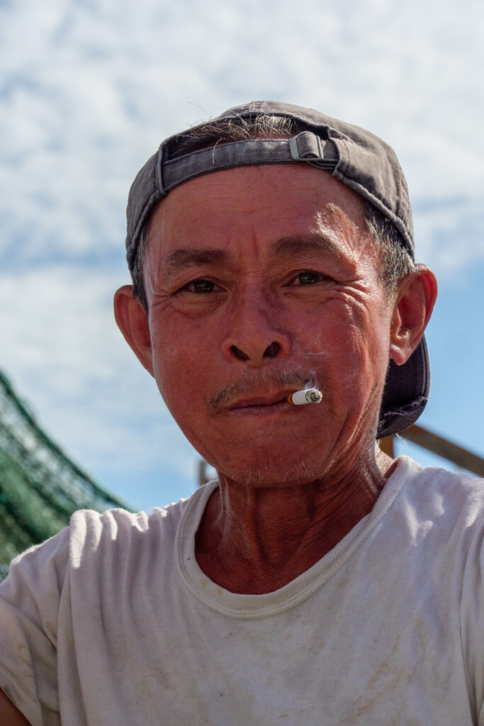 An older Vietnamese man smokes a cigarette, behind him is a hanging shrimp net on his boat, and the ocean in the sun. He's wearing a backwards, weathered blue ballcap.