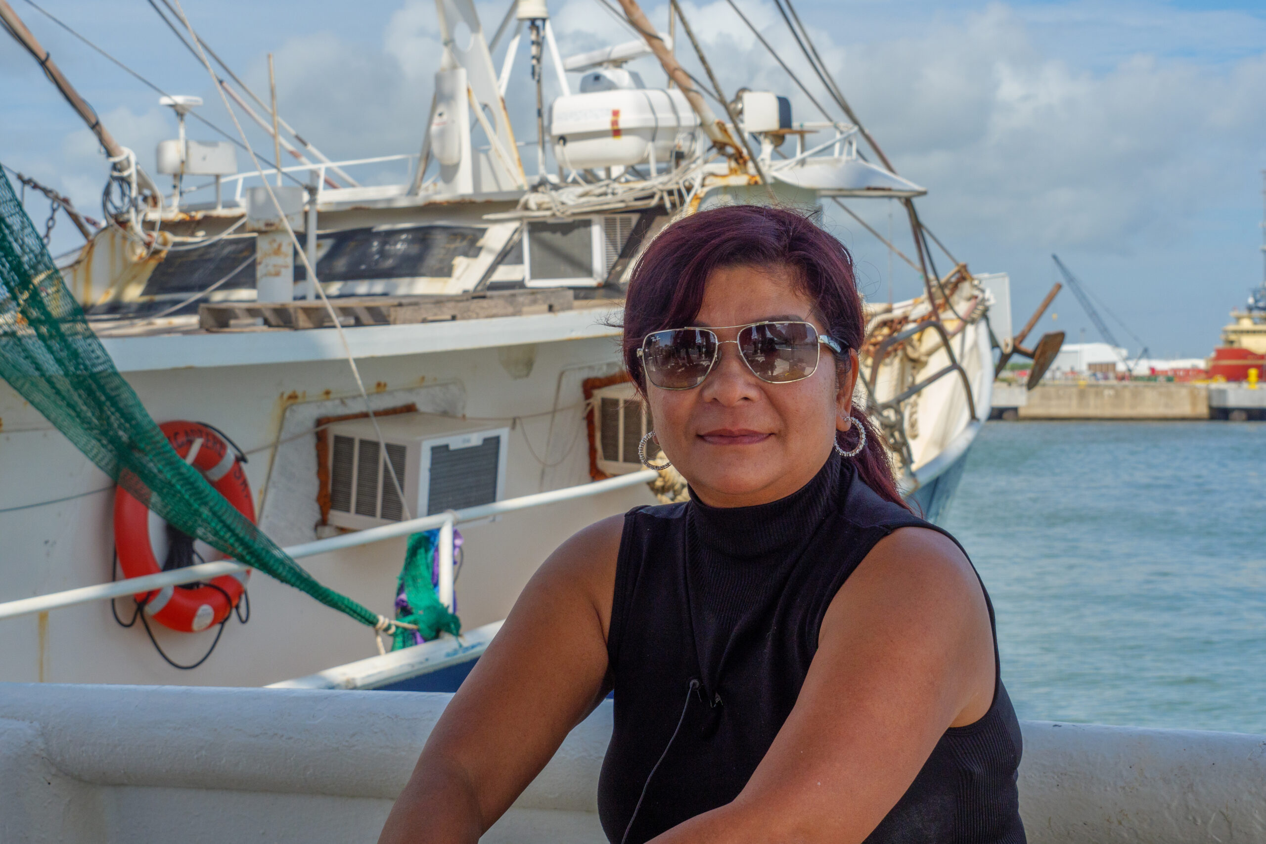 A Vietnamese in sunglasses with short black hair looks at the camera, a shrimp boat floating in the background behind her under a partly cloudy sky.