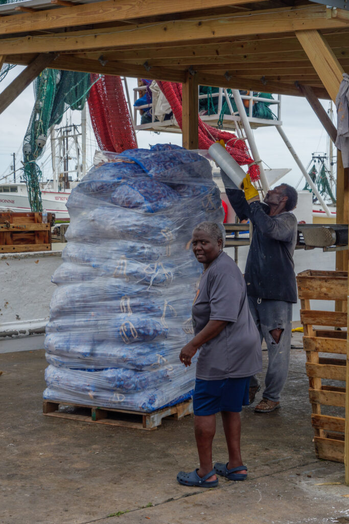 Two Black dockworkers wrap a huge pile of bags of shrimp on top of a pallet in plastic wrap.