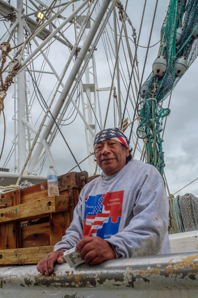 A Hispanic man poses by the edge of a fishing boat, with rigging and nets hanging behind him. His hands are at the railing, with a pack of Marlboro cigarettes held in one.