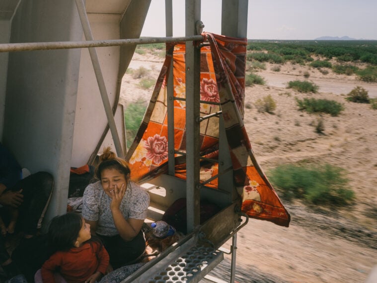 The mother and child are nestled into a thin area of relative "safety" at the back of the train car, with a thin orange and yellow shawl or tapestry tied behind them to shade their skin from the rays of the sun.