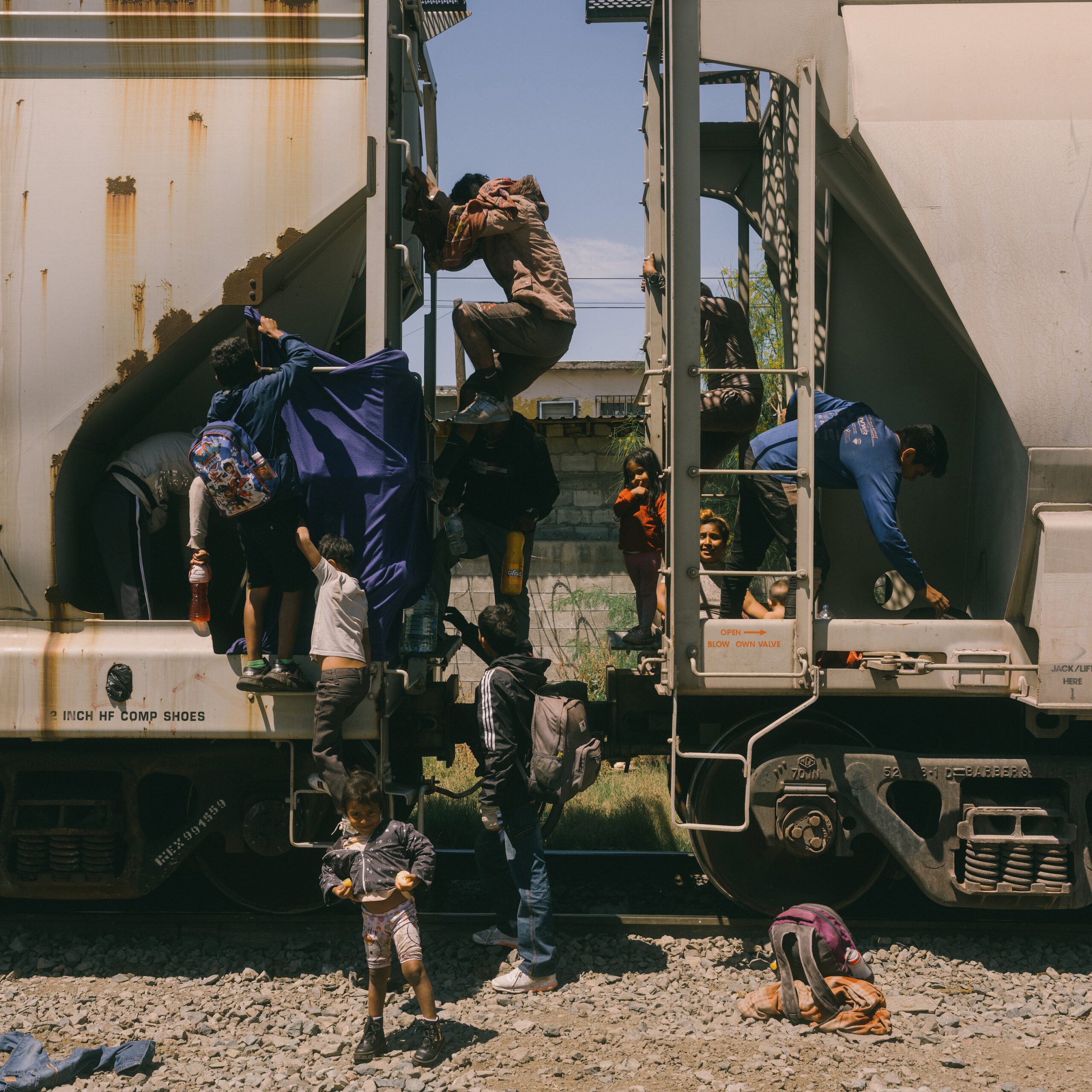 A pair of families photographed in the process of disembarking from their stowaway hiding places aboard La Bestia. A young child stands proudly on the ground while other family members climb or cling to the sides of the train.
