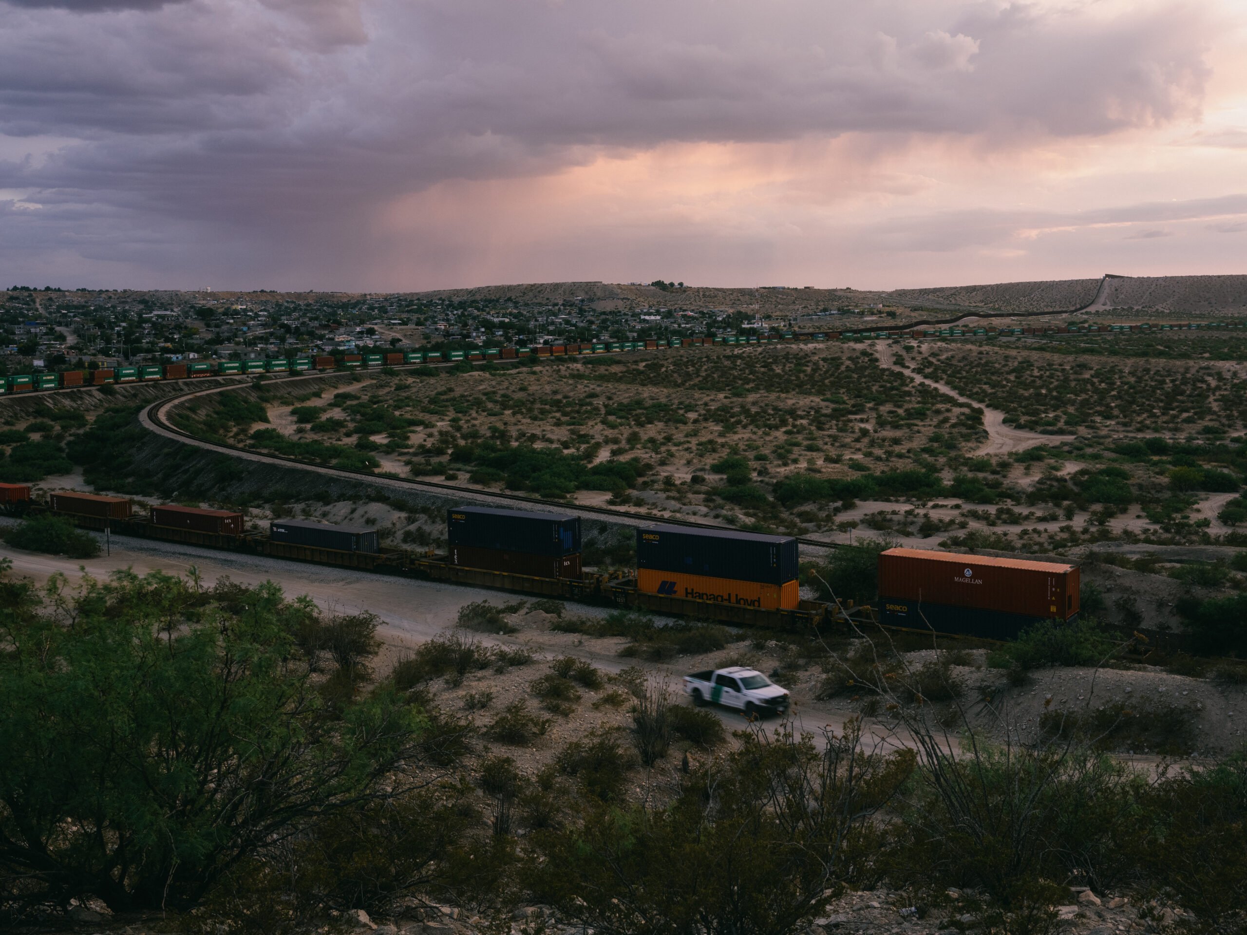 A wide-angle view of the train curving through the countryside. The border patrol pickup truck seems tiny in comparison to the massive train. The sky is cloudy and ominous.