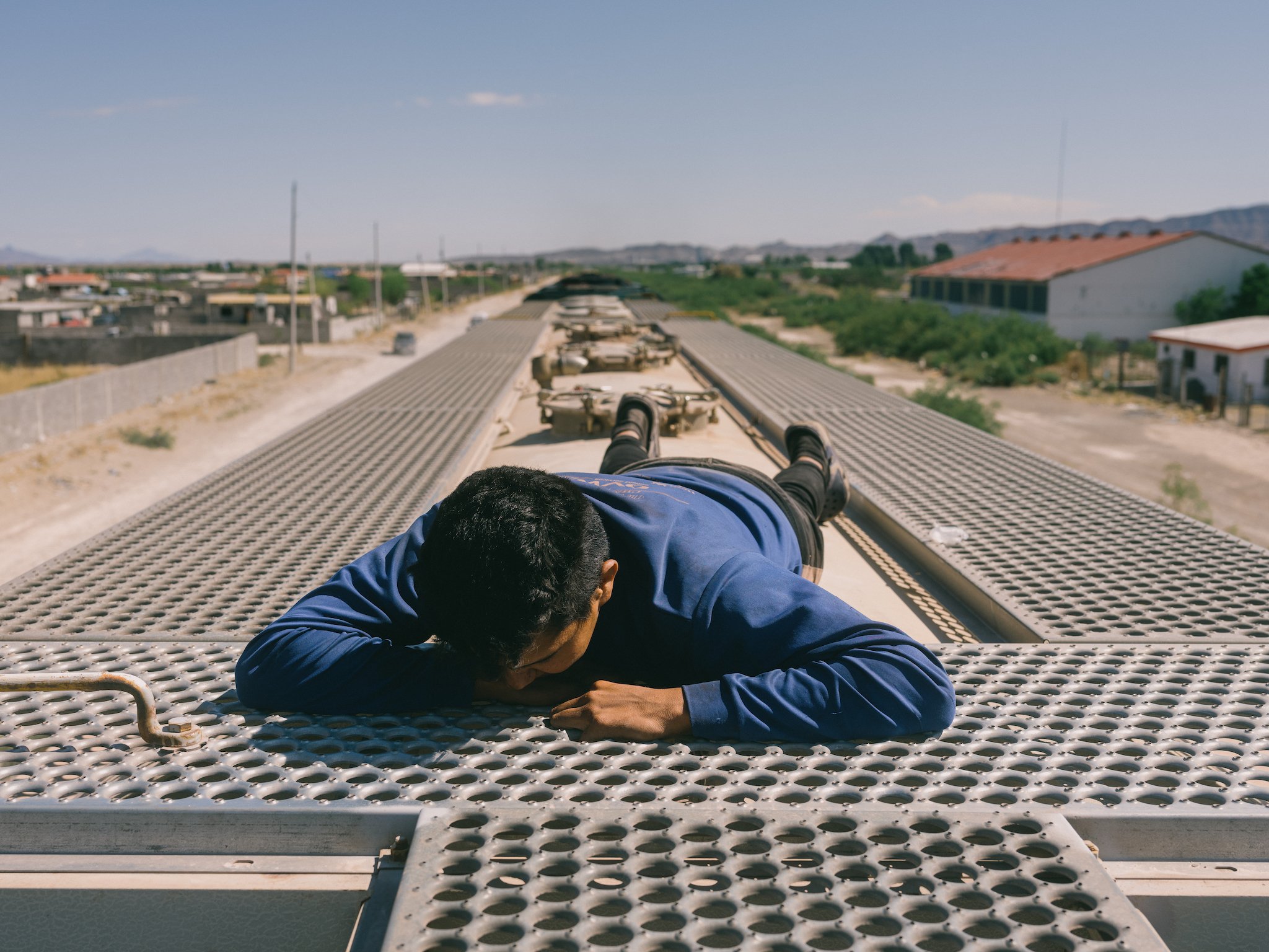 A man lies prone, face down, on the top of a train as it moves through the desert under a clear sky.