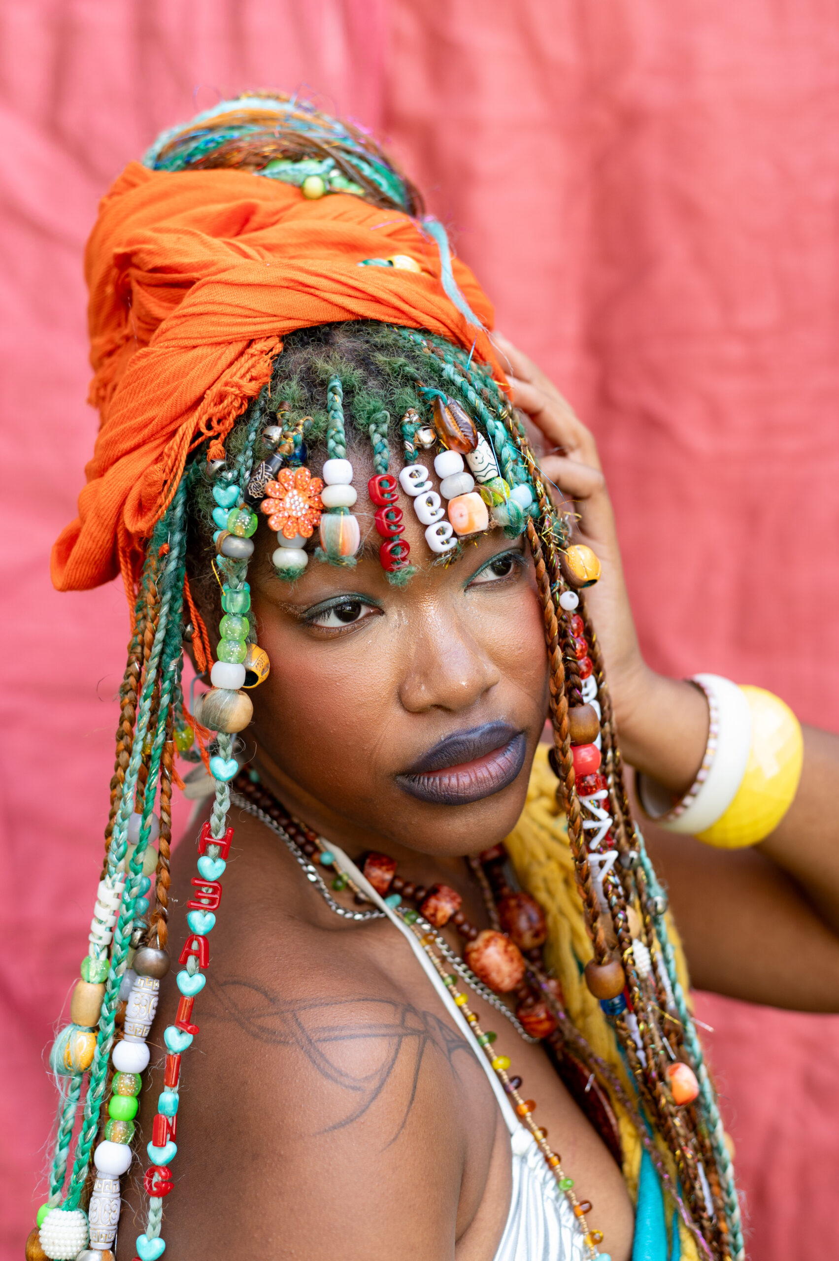 A Black woman stands in profile, looking over her right shoulder towards the photographer's right. She is wearing an orange silk scarf over her elaborately beaded locks.