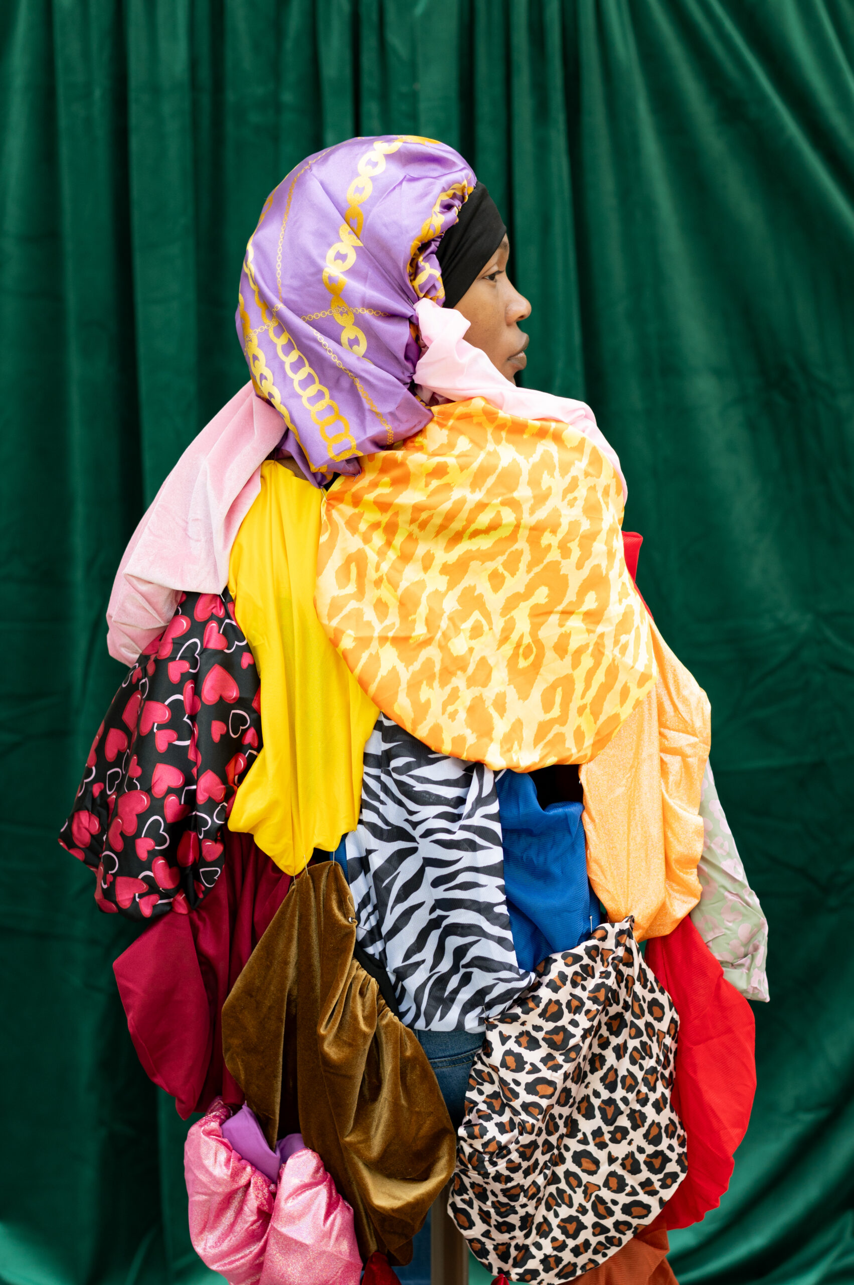 A Black woman stands in profile, her full-body length locks covered in an elaborate wrap made from multiple, colorful patterned soft looking fabrics.