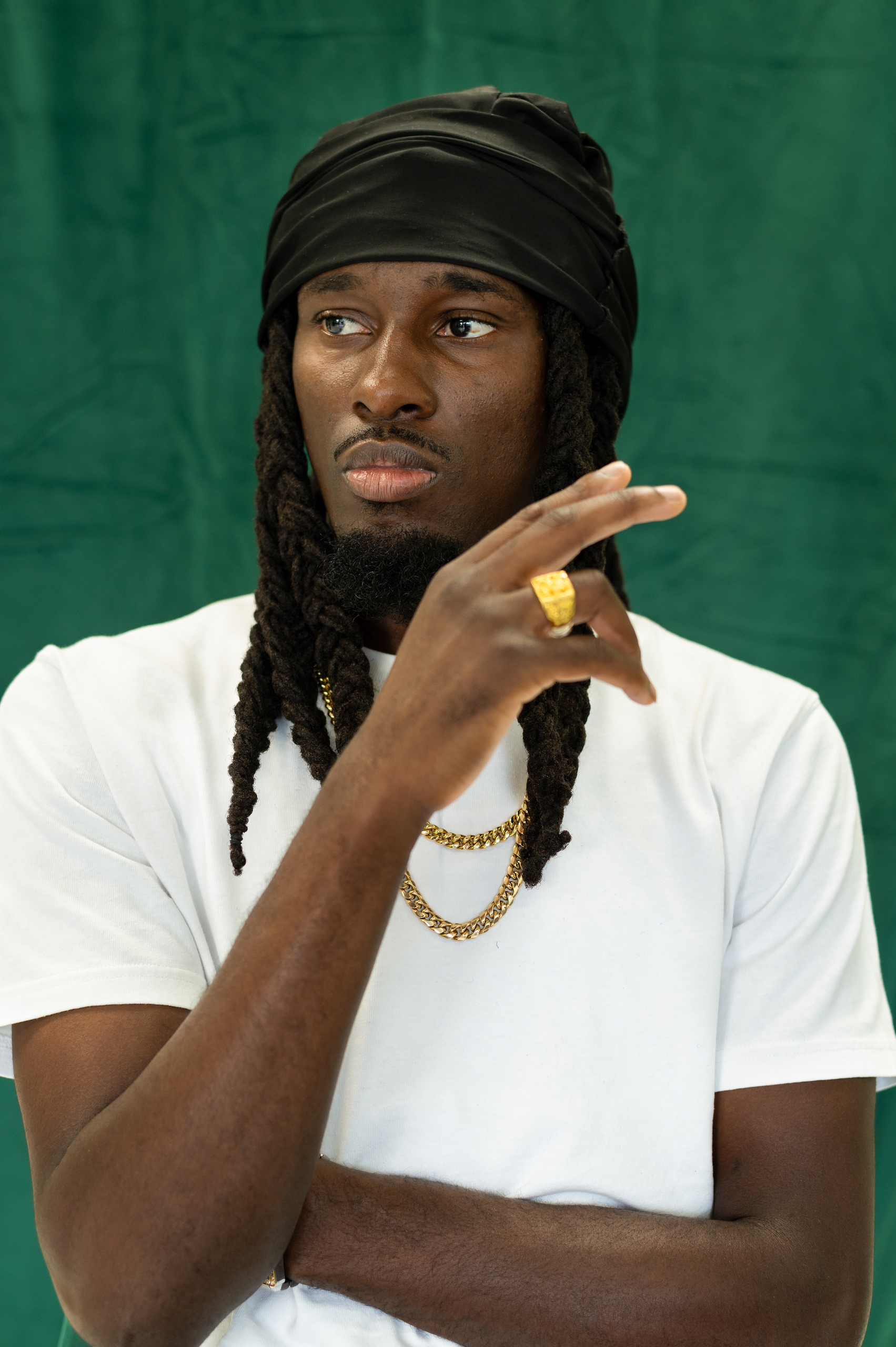 A Black man with thick locks spilling from under a black silk scarf on his head. He is wearing a white tee with gold chains, and is gesturing with his hand decorated with a large golden ring.