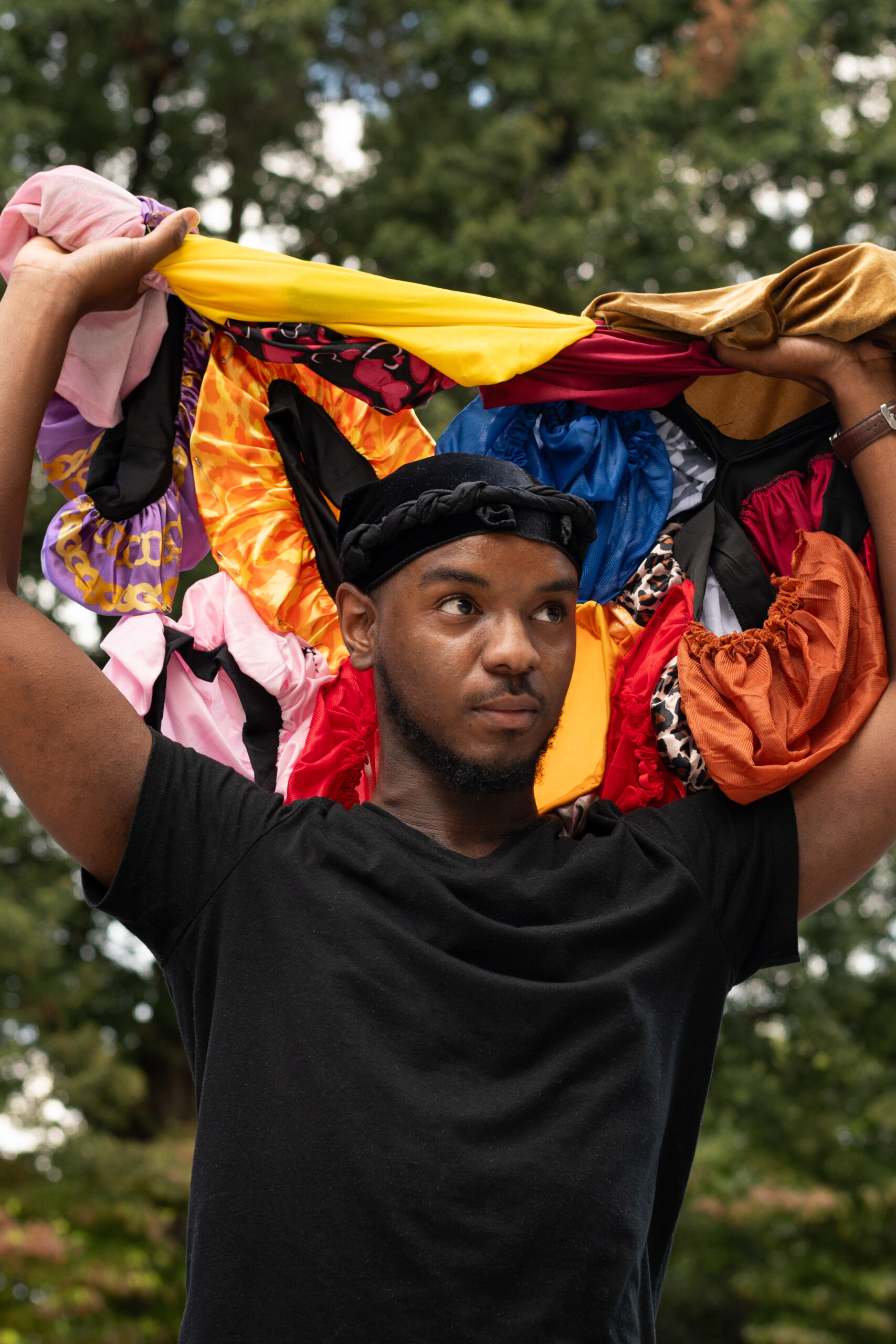 An outdoor portrait of a Black man, looking slightly to the right of the photographer. He is holding up his ornate head covering, made from several silky or satiny fabrics of different colors bright colors.