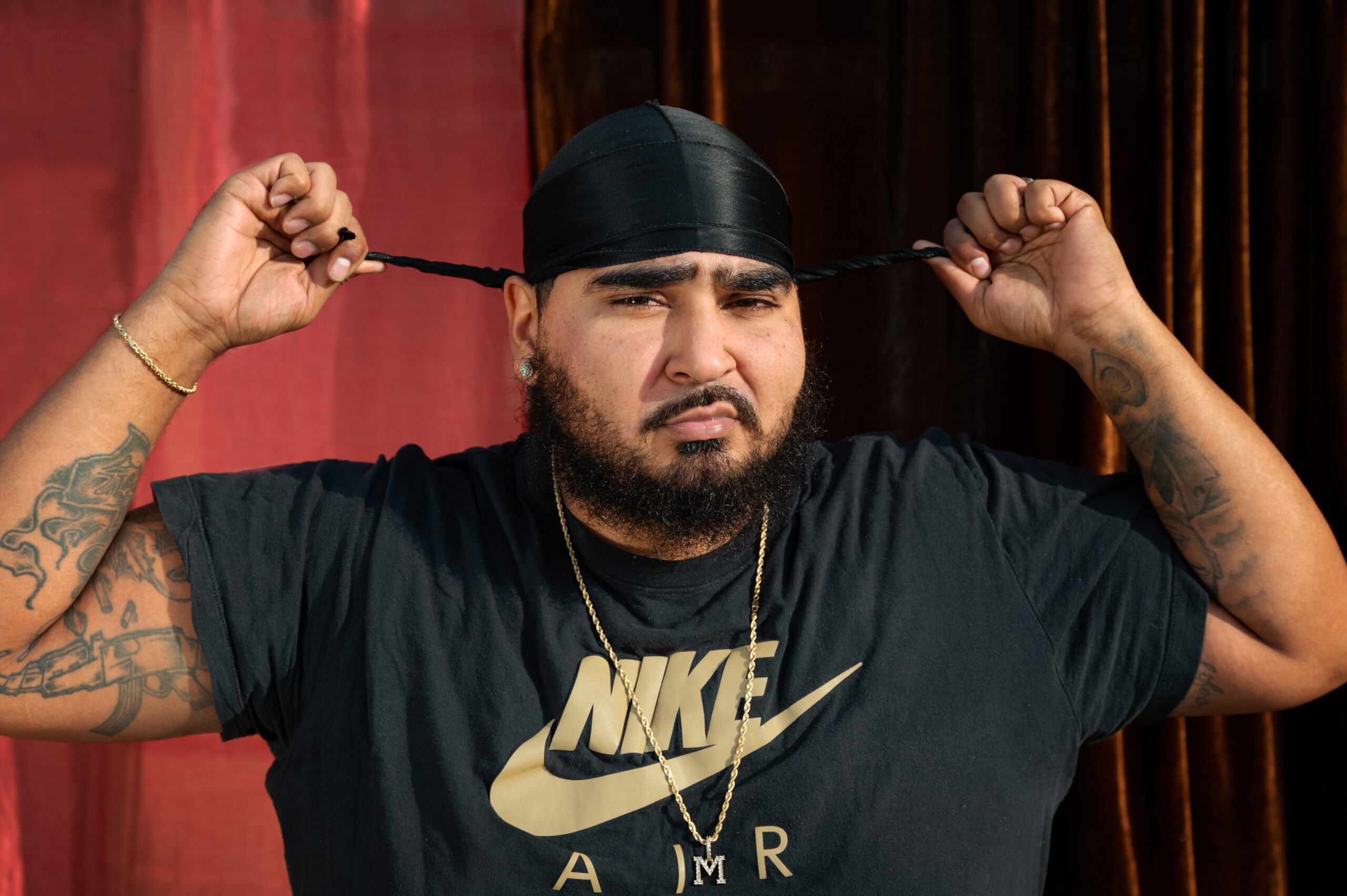 A Black man with a bushy black beard and visible tattoos under his NIKE Air t-shirt adjusts his black satin hair wrap.