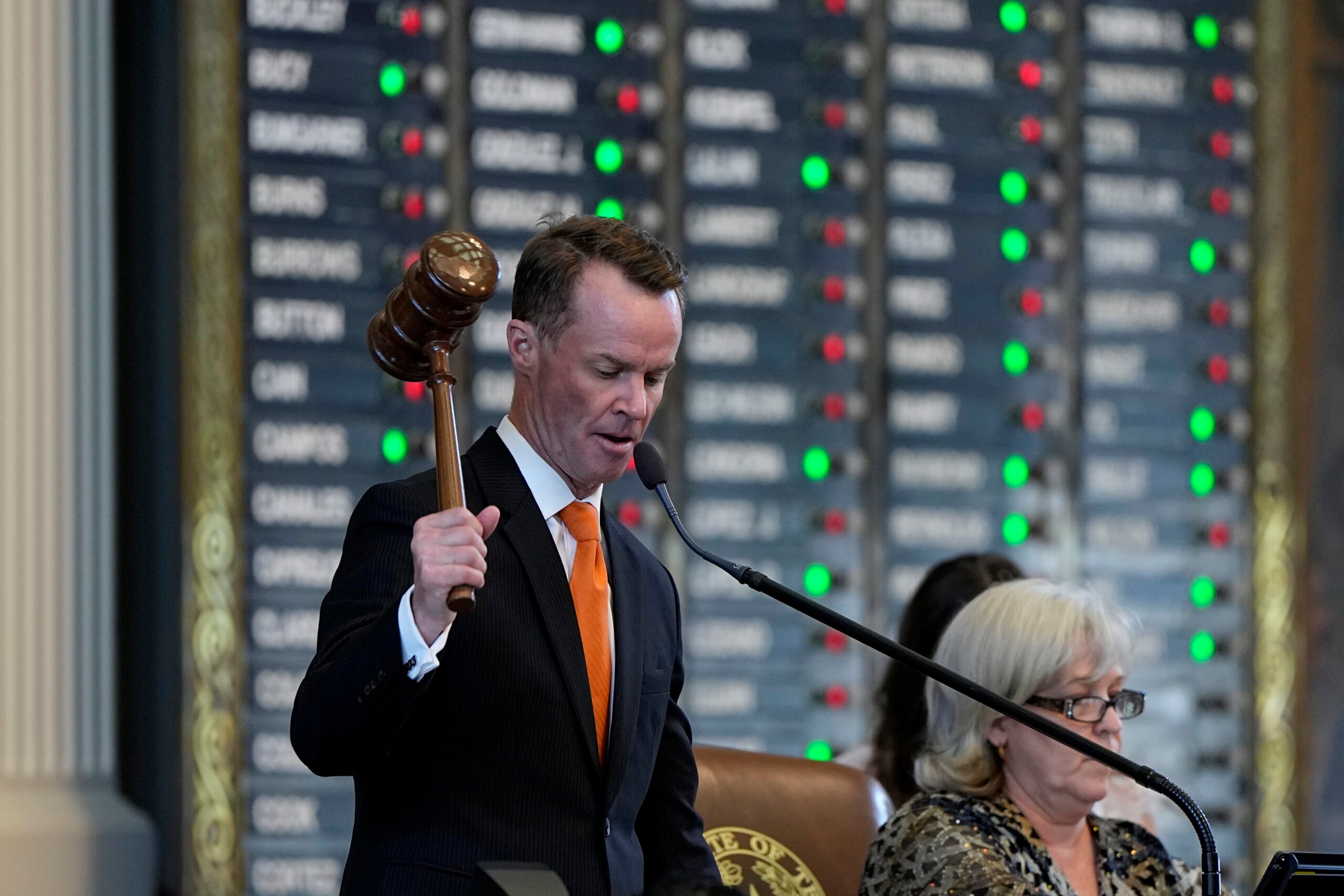 Dade Phelan, a white man in a suit and tie, prepares to bang his gavel. Behind him is a light board showing how different representatives vote on the current bill.