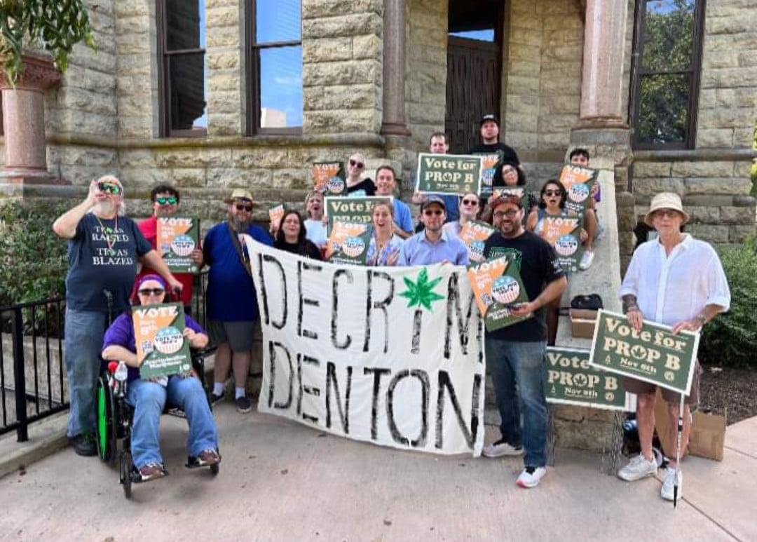 A diverse group of a few dozen people gather with a large "Decrim Denton" banner" and signs supporting reform of marijuana laws.