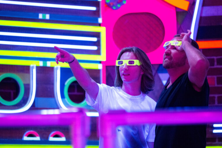 Two visitors to Meow Wolf Grapevine stand among the neon signs at the Neon Kingdom, one pointing as she looks through paper refractive glasses and the other lifts his glasses to look at what she's pointing at.