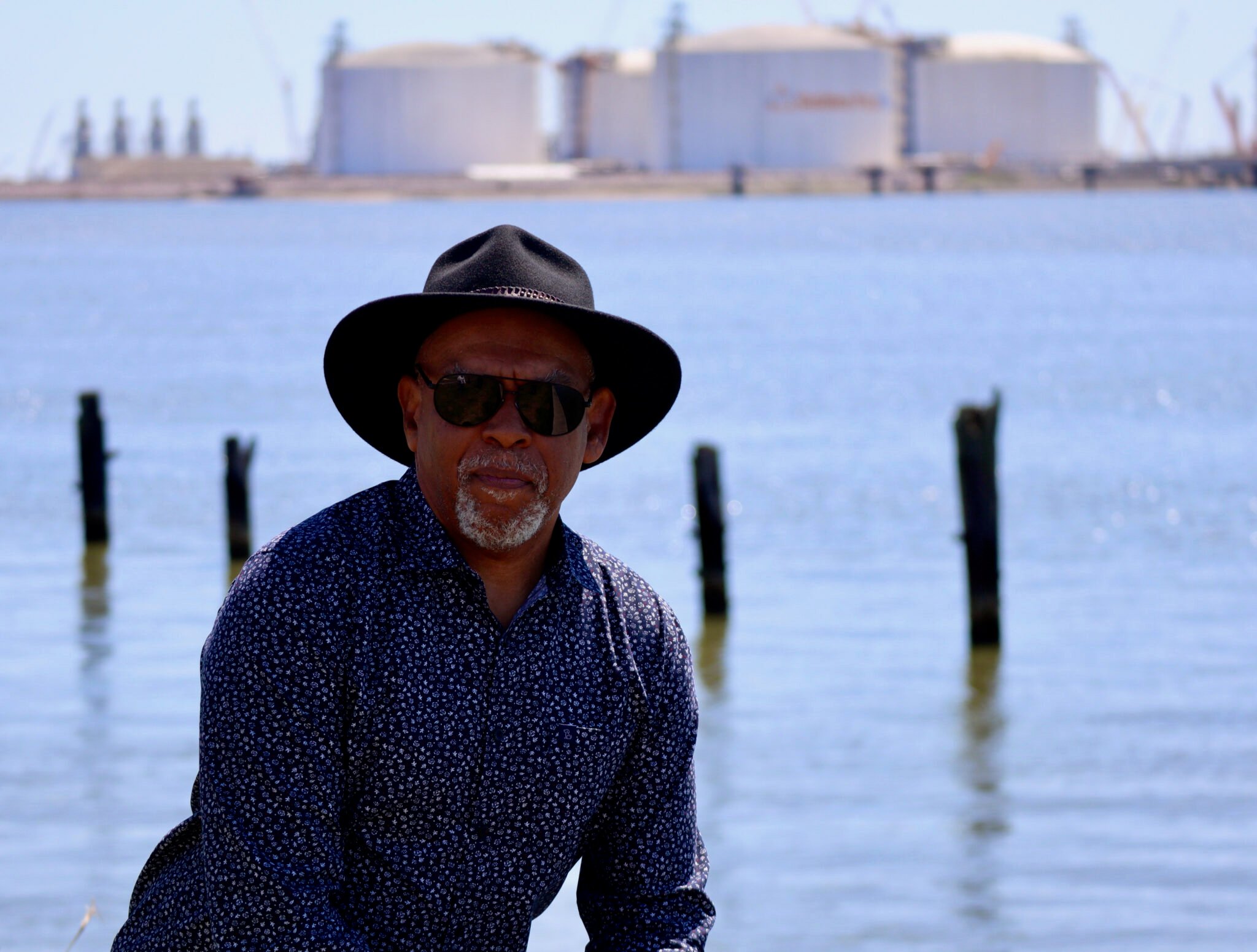 A Black man in a black panama hat and dark blue dot-patterned shirt poses by the water with an LNG terminal behind him.
