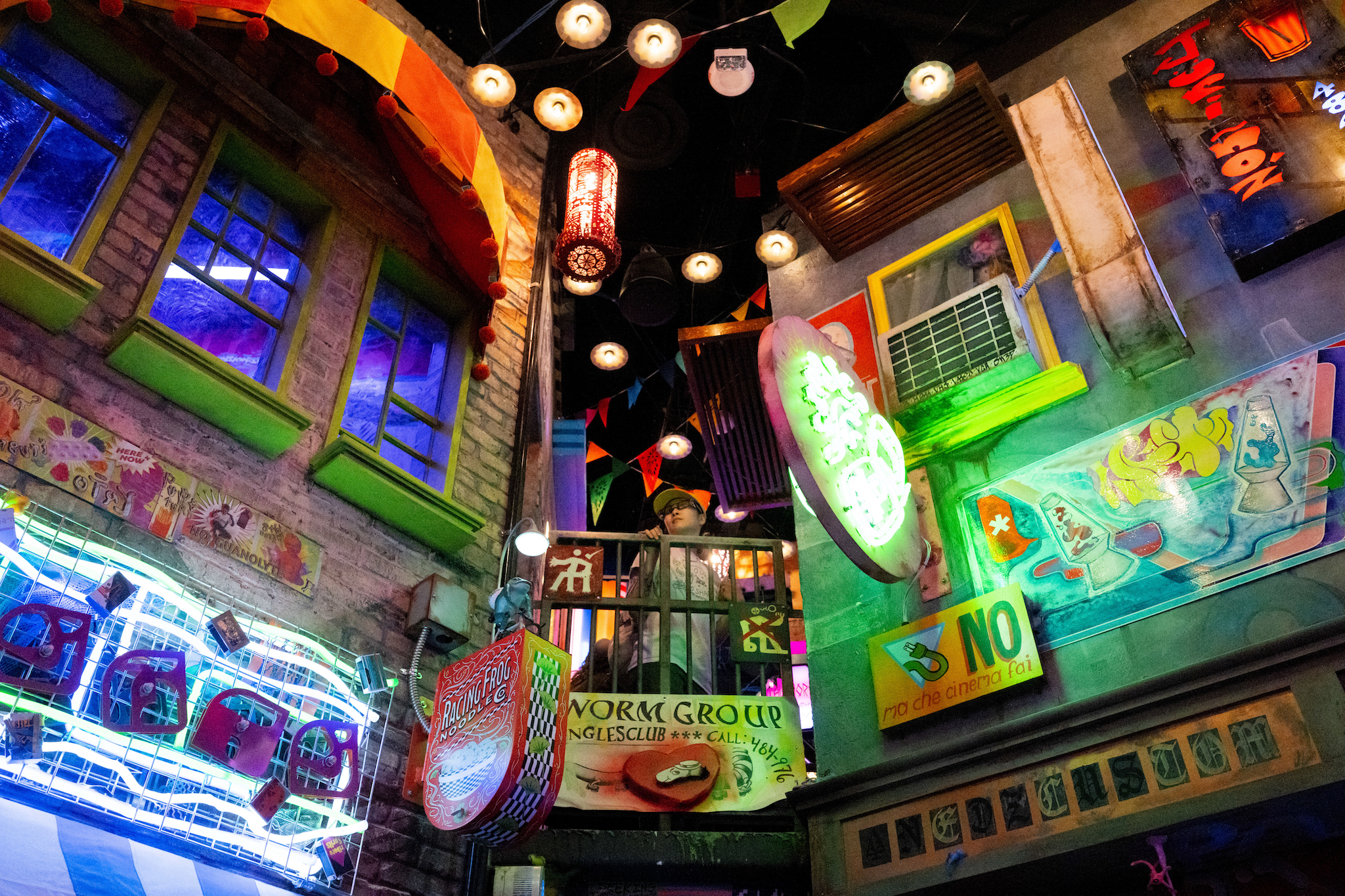 A visitor pauses, holding the railing as they look down into the art installation. The walls are dotted with commercial advertisements and signs for imaginary shops, many in made up or alien languages. One reads Worm Group Englesclub, the print dripping off at a weird curve.