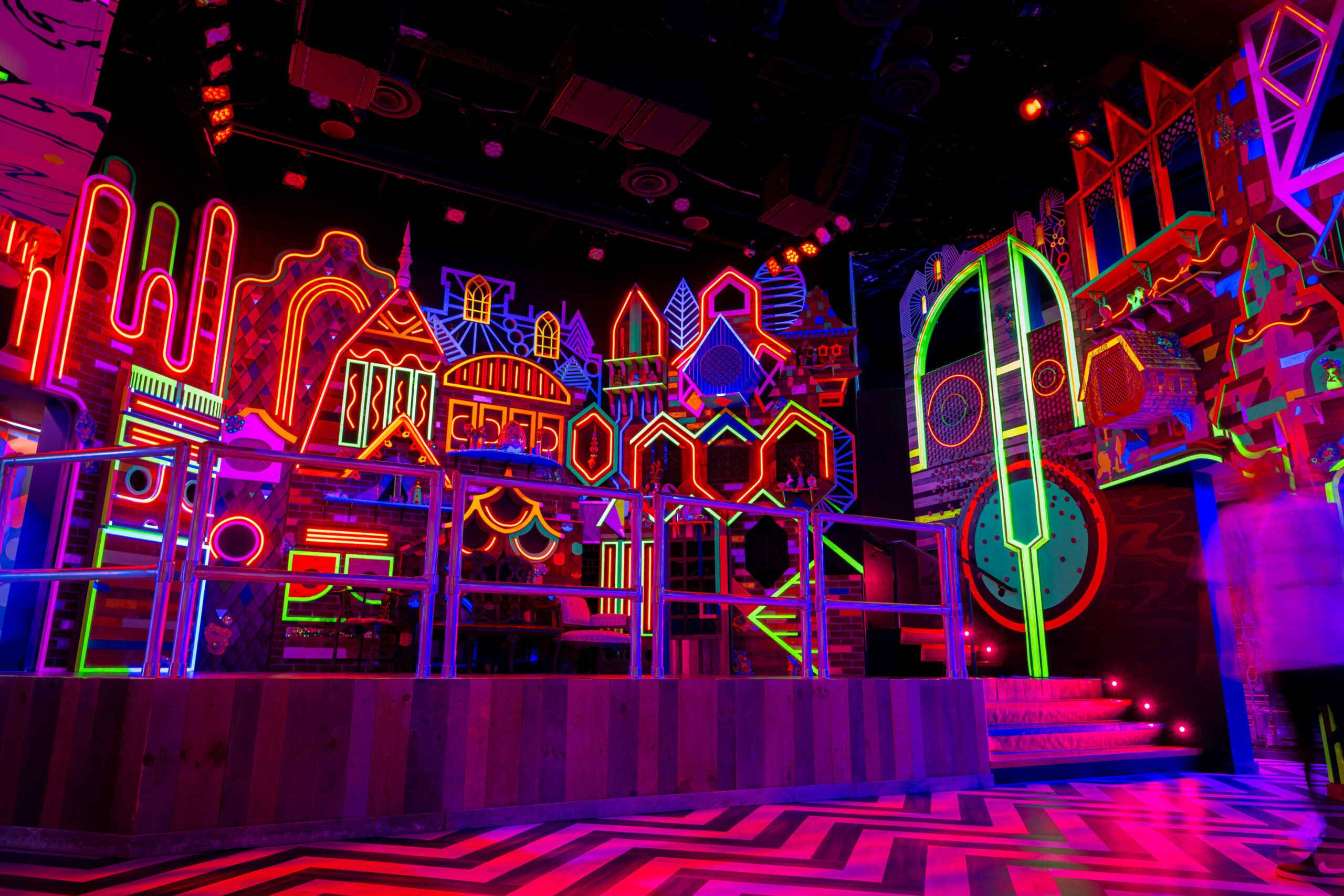 A stage with a backdrop of neon lights made to look like. fantasy walled cityscape in glowing rainbow lights.