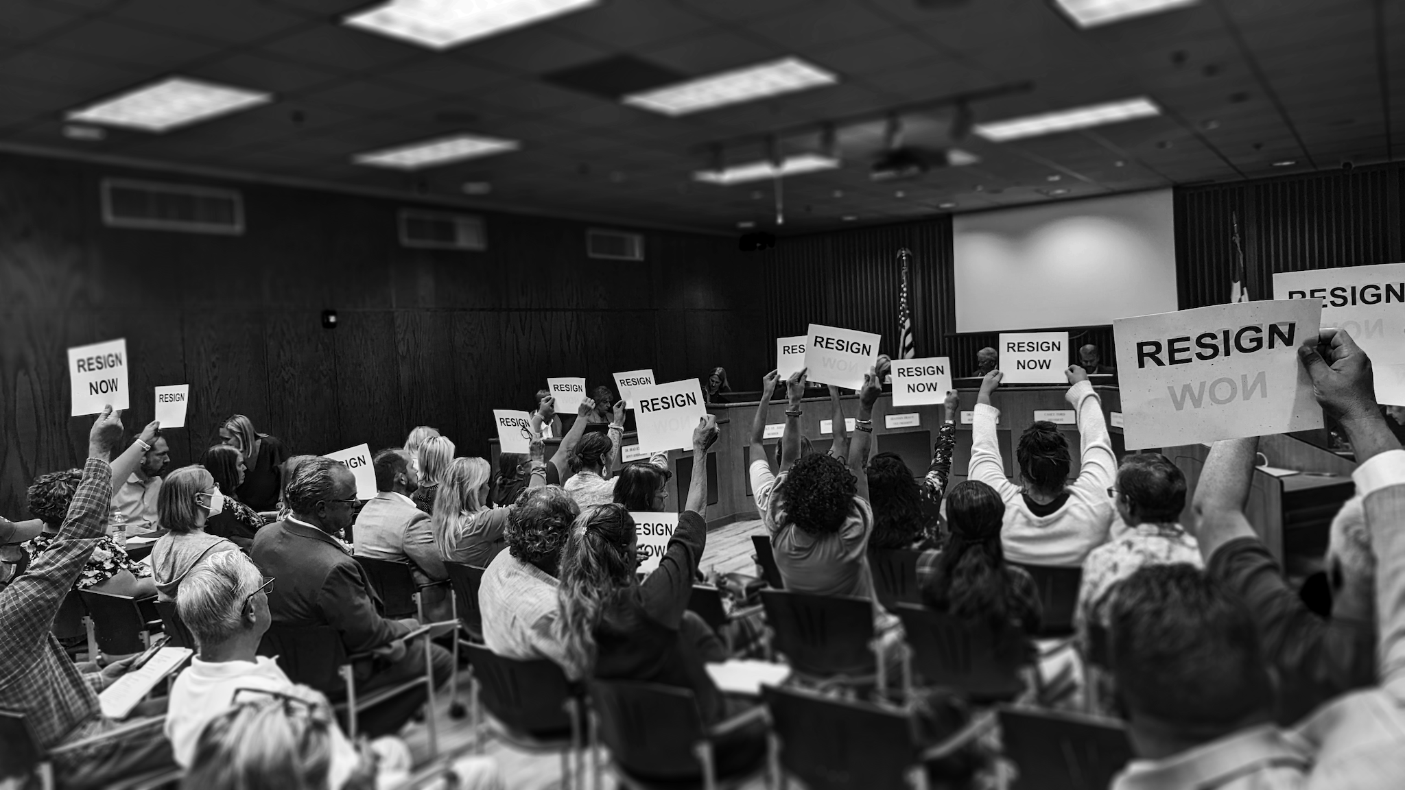 In a black and white photo, audience members hold up Resign Now signs in the board room of a school board.