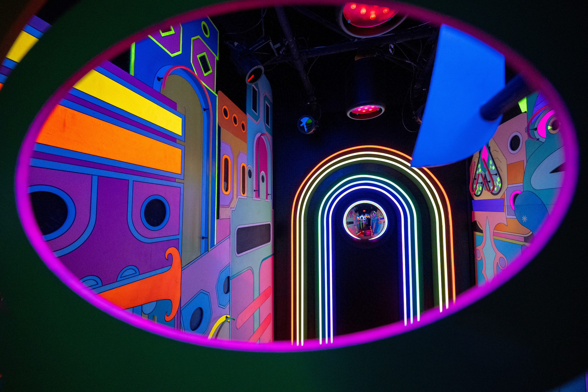 An oval shaped window peeks into a brightly lit hallway with neon lights and colorful paintings glowing on the walls.