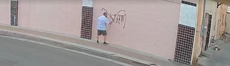 Surveillance footage shows a man in a checkered, light blue shirt spray-painting "No Islam" on an exterior wall with an "X" over it.