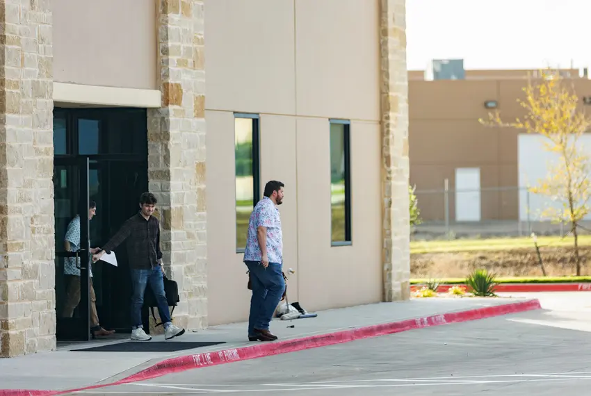 A photograph taken at a medium distance in a parking lot of three people leaving an GOP office building in Texas on a sunny day. White supremacy extremist Nick Fuentes is caught in the middle, in dark clothes with a frown for the photographer.
