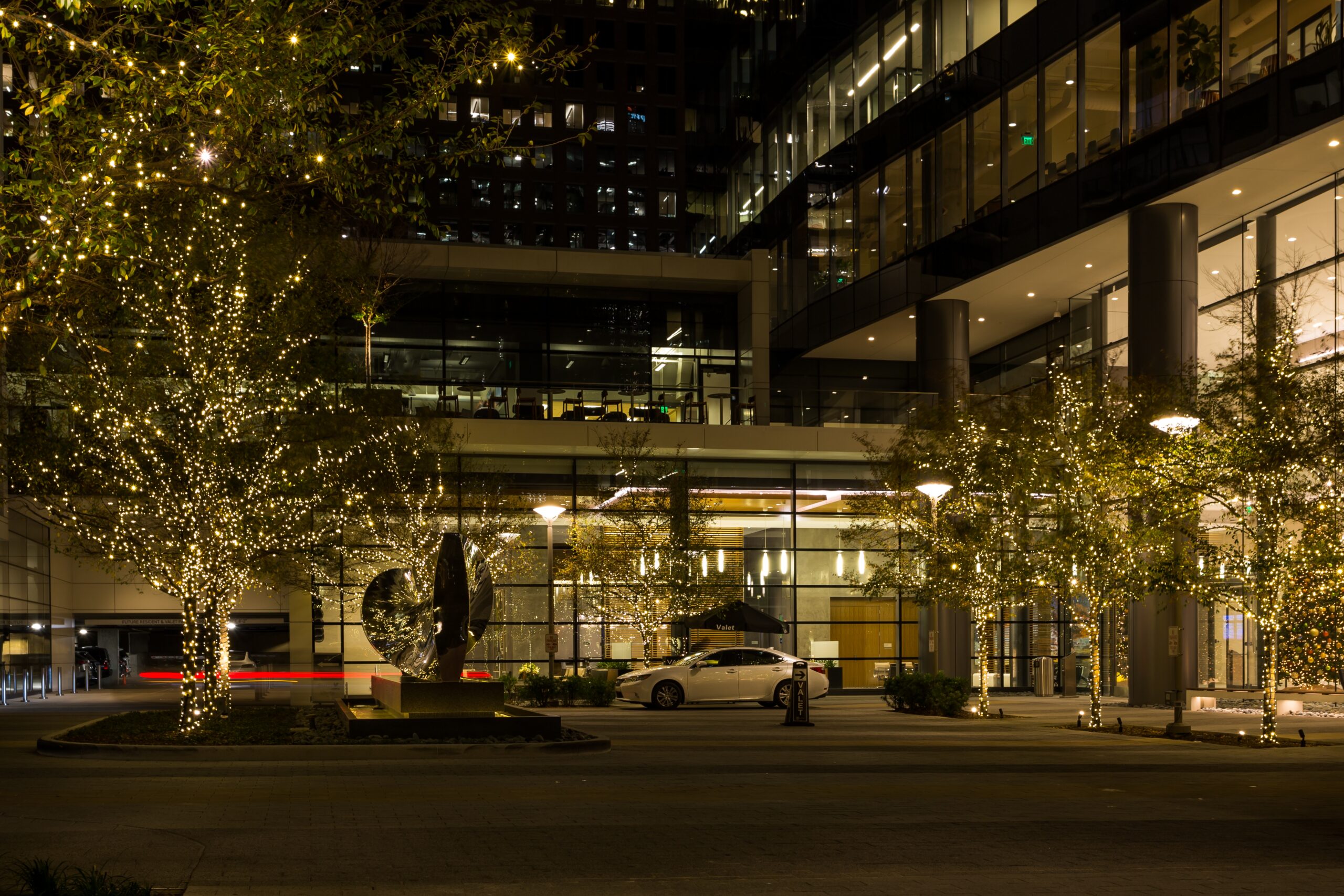 A fancy car is parked in front of a luxury apartment building, all glass windows, with Christmas lights in the trees. The effect is festive but sterile.