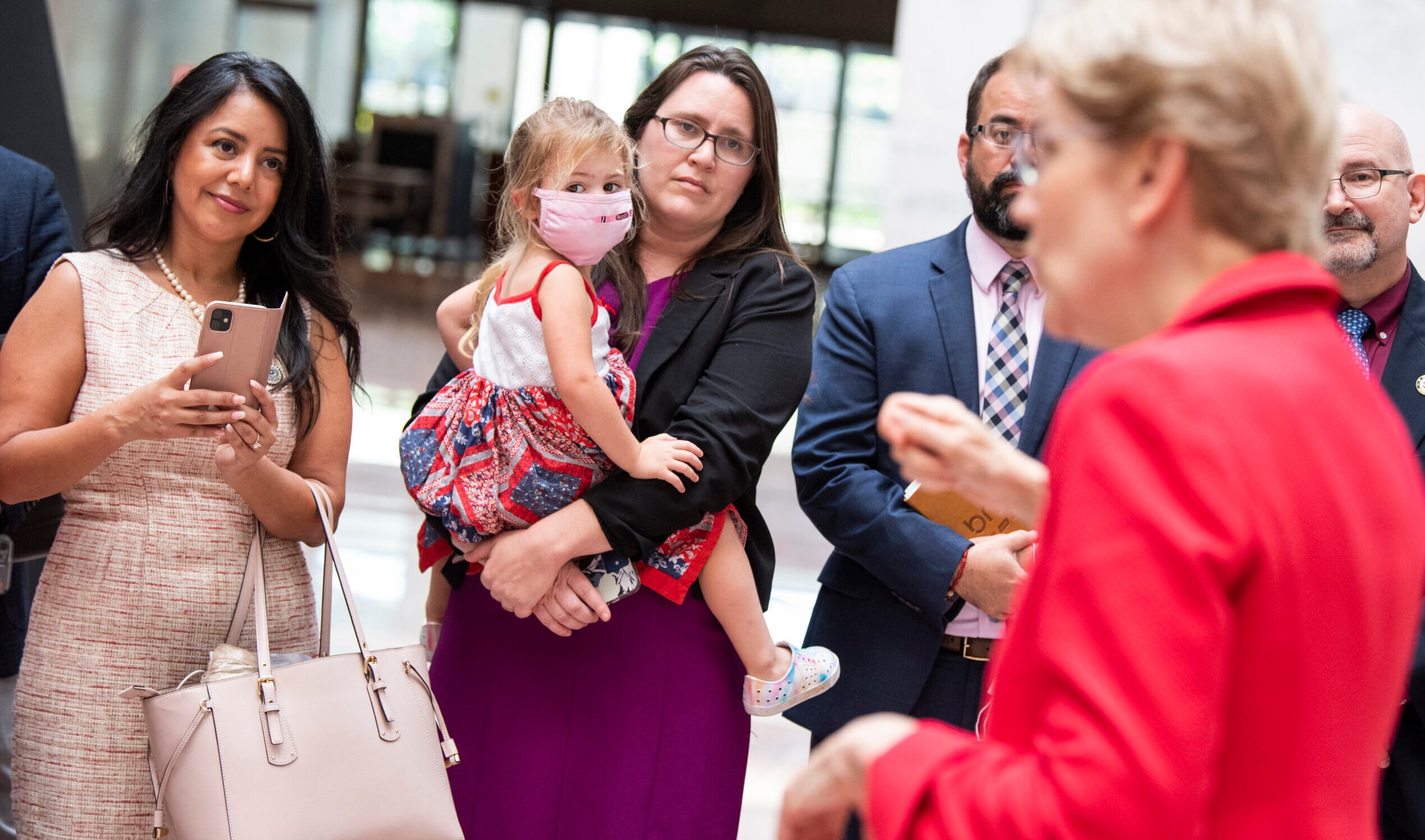 Erin Zwiener, a white woman with brown hair, holds her toddler daughter, wearing a mask, as she stands with other Democrats listening to Elizabeth Warren, who is wearing a red suit jacket and out of focus in the foreground.