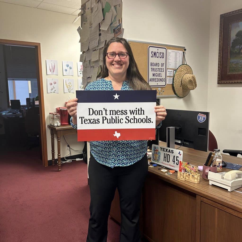Erin Zwiener, a white woman with long brown hair and red plastic glasses, poses with a Don't Mess with Texas Public Schools sign in her office.