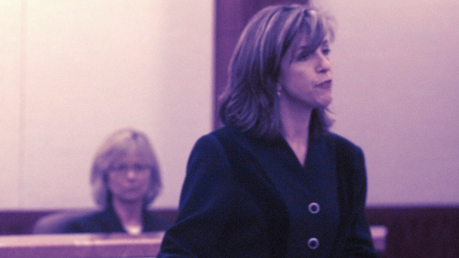 A photo of Kelly Siegler, a blonde woman, in a dark suit jacket, in a courtroom. An amethyst color filter was added to the photo to give it a colder feeling.