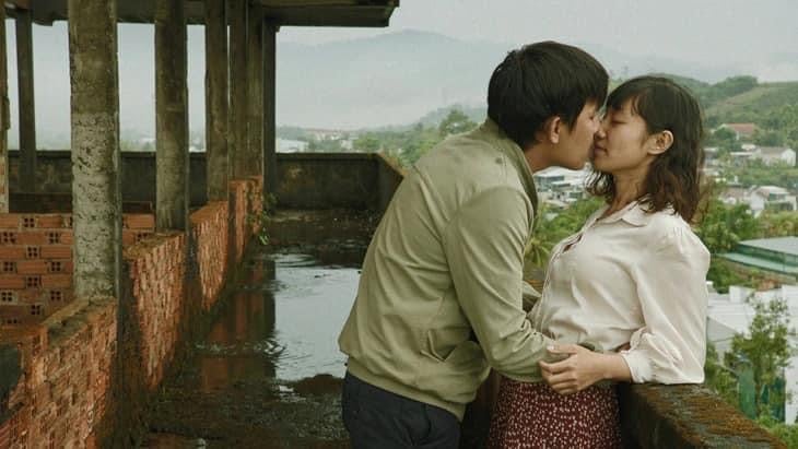 A Vietnamese man in a tan jacket embraces a woman in a white blouse and red patterned skirt, about to kiss her as they stand together on a rain-filled terrace balcony of a brick building.