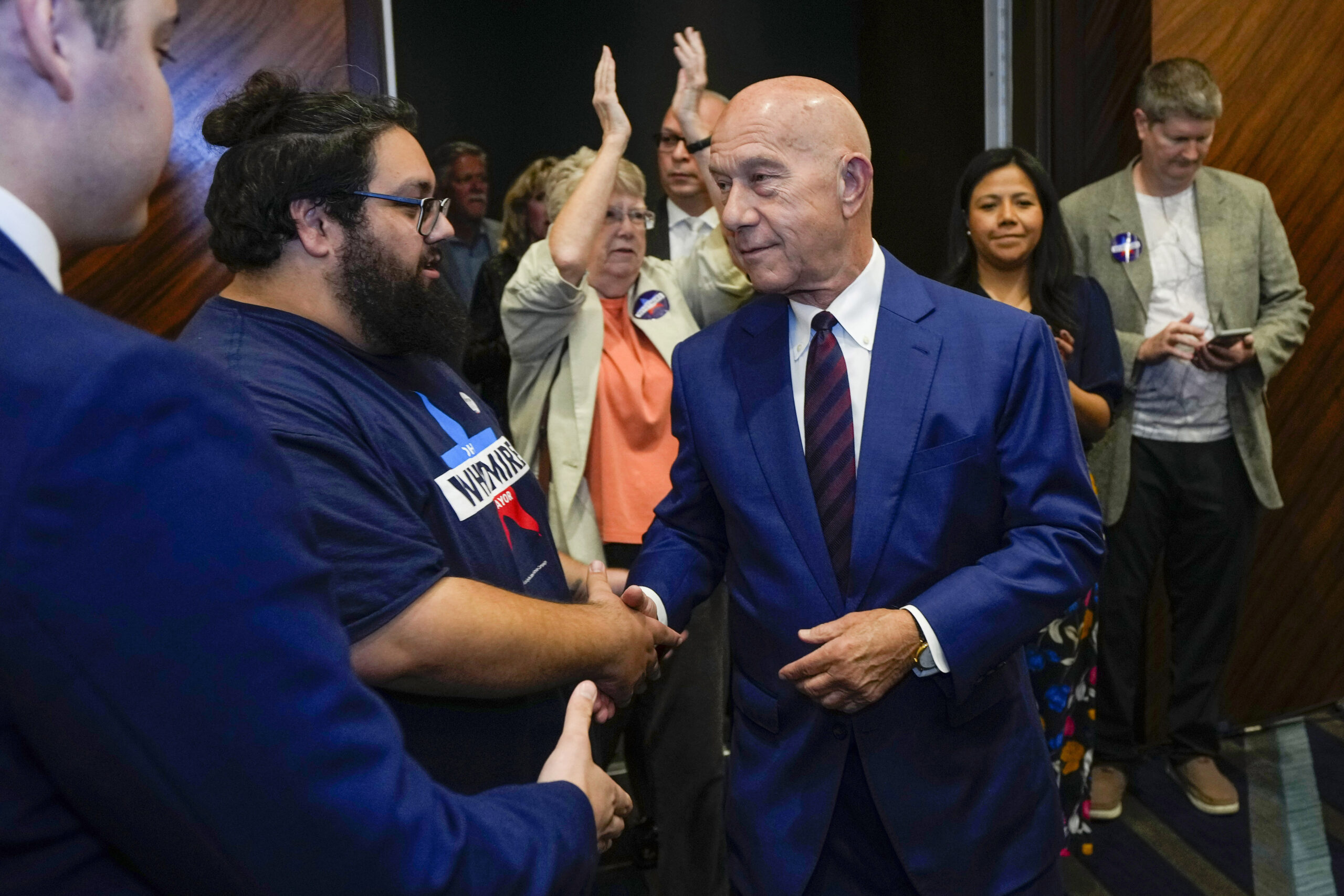 State Sen. John Whitmire, a bald white man, shakes hands with a bearded supporter at crowded public event. Whitmire is wearing a blue suit and red tie.