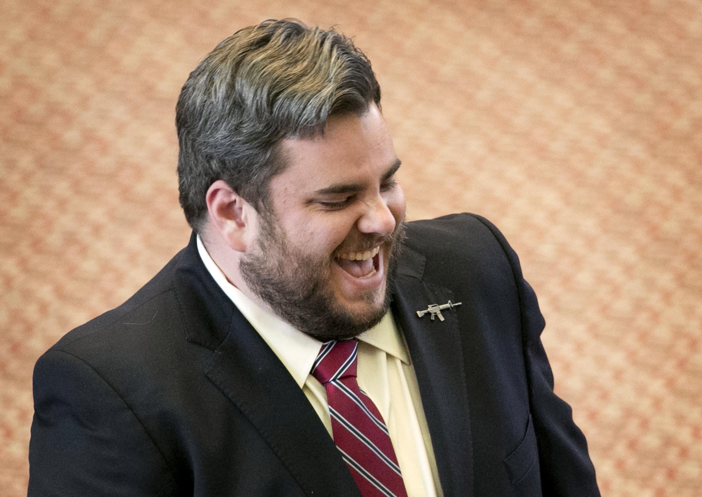 Jonathan Stickland, a white man with a light beard and mustache, wearing a suit with a striped red tie, laughs in a candid portrait photo.