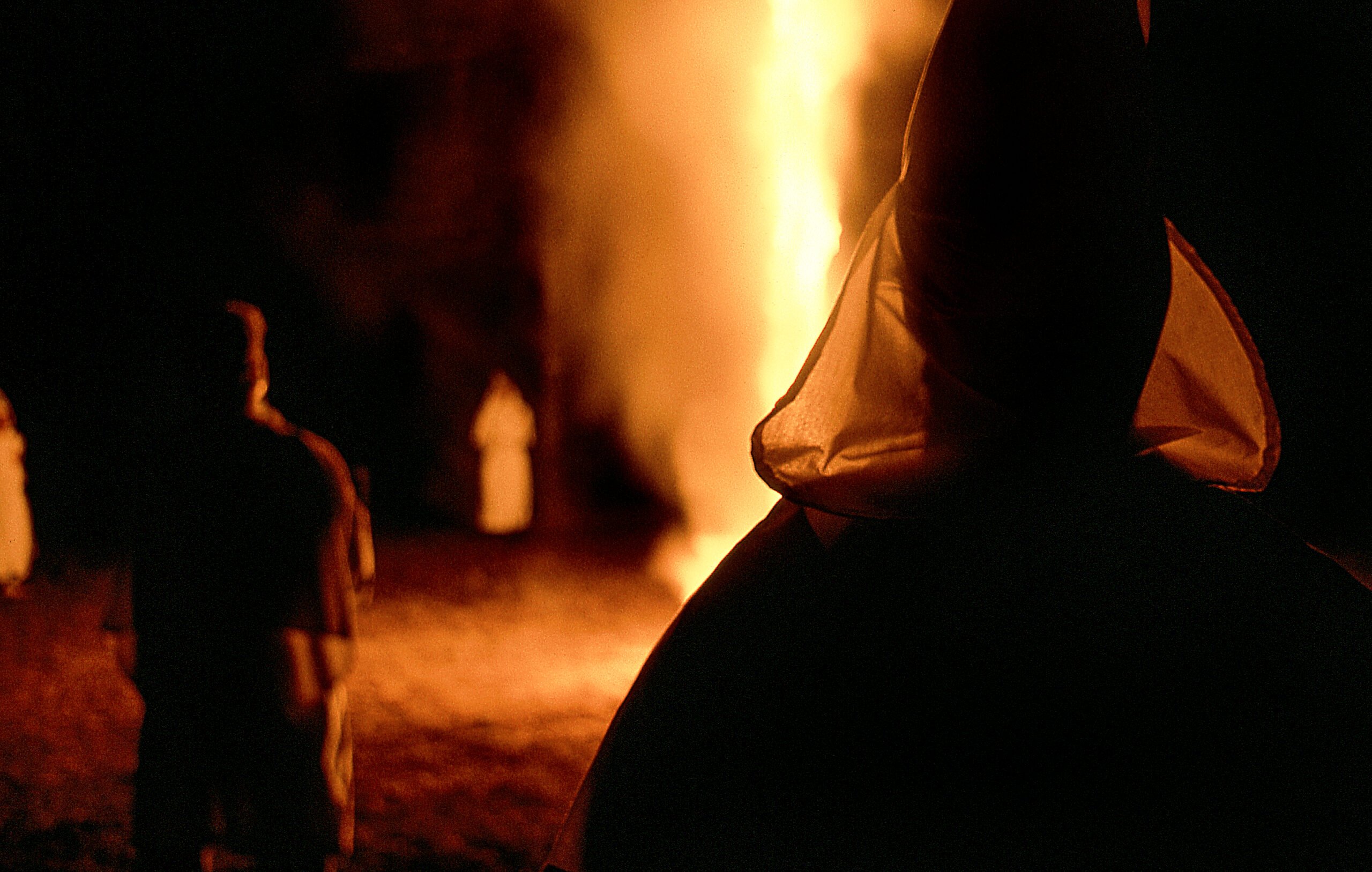 A shadowy photo of Ku Klux Klan members, silhouetted in their white robes by a burning bonfire.